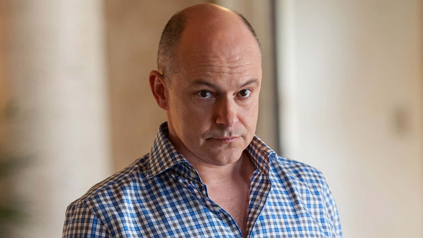 Rob Corddry 桌布 壁紙 — 一位光頭男子身穿藍色格紋襯衫，在室內以嚴肅的表情專注地凝視。
