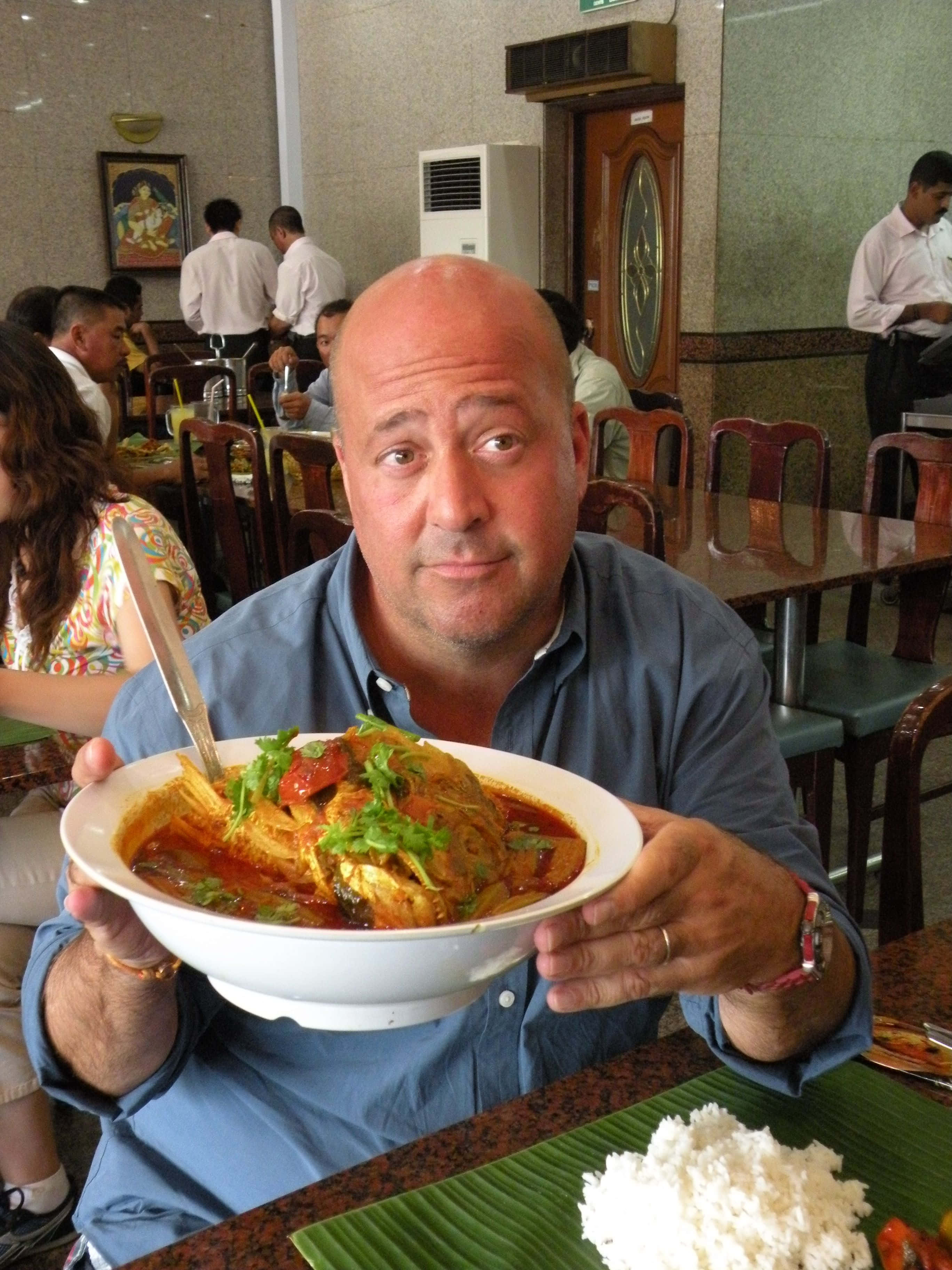 Food Photography 桌布 壁紙 — 一位光頭男子身穿藍色襯衫，在繁忙的餐廳中自豪地展示一碗冒著熱氣、點綴著新鮮香菜的濃郁咖哩。