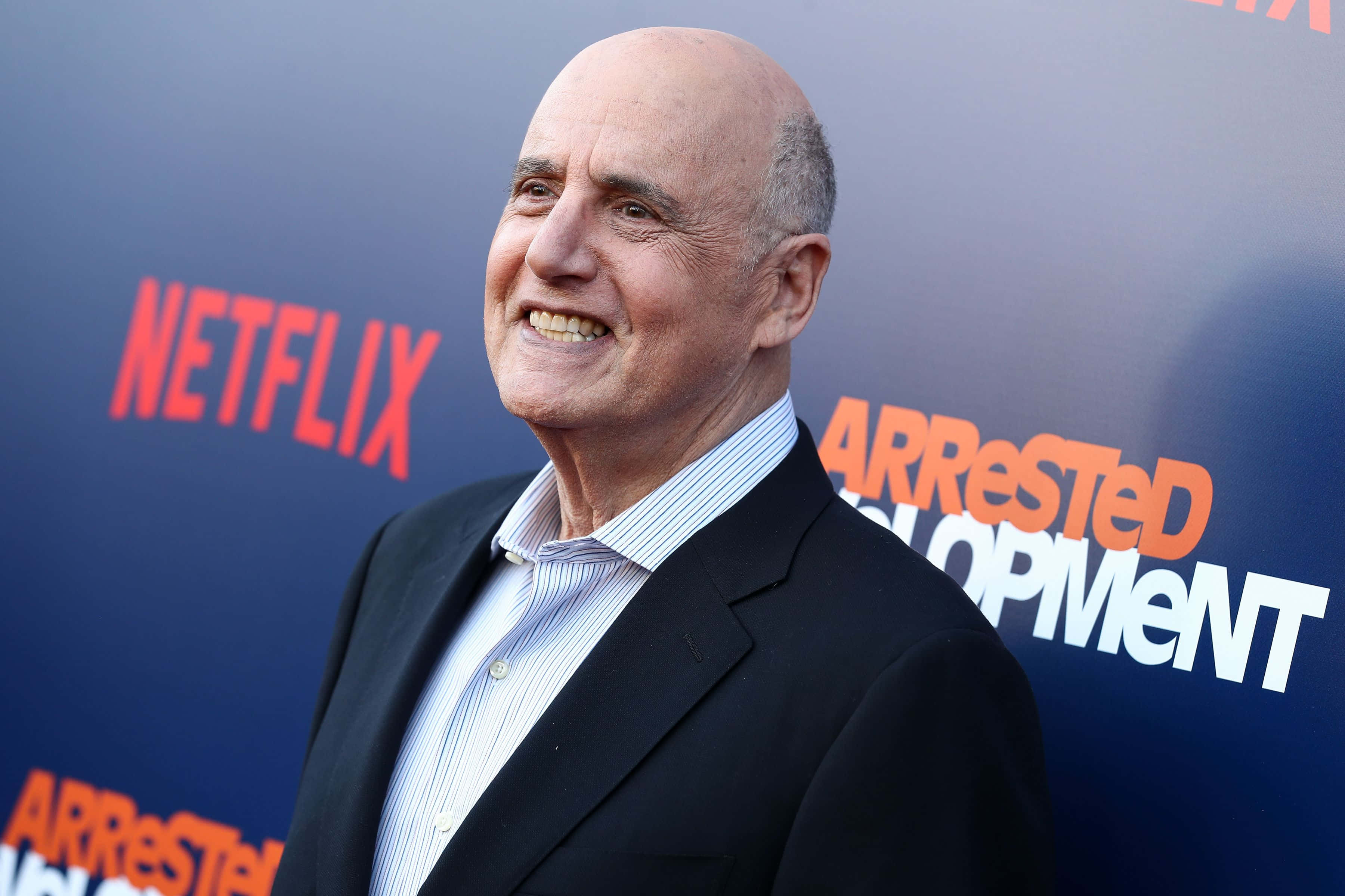jeffrey tambor 桌布 壁紙 — 一位光頭男子身穿黑色西裝，在Netflix《Arrested Development》活動背景前燦爛微笑。