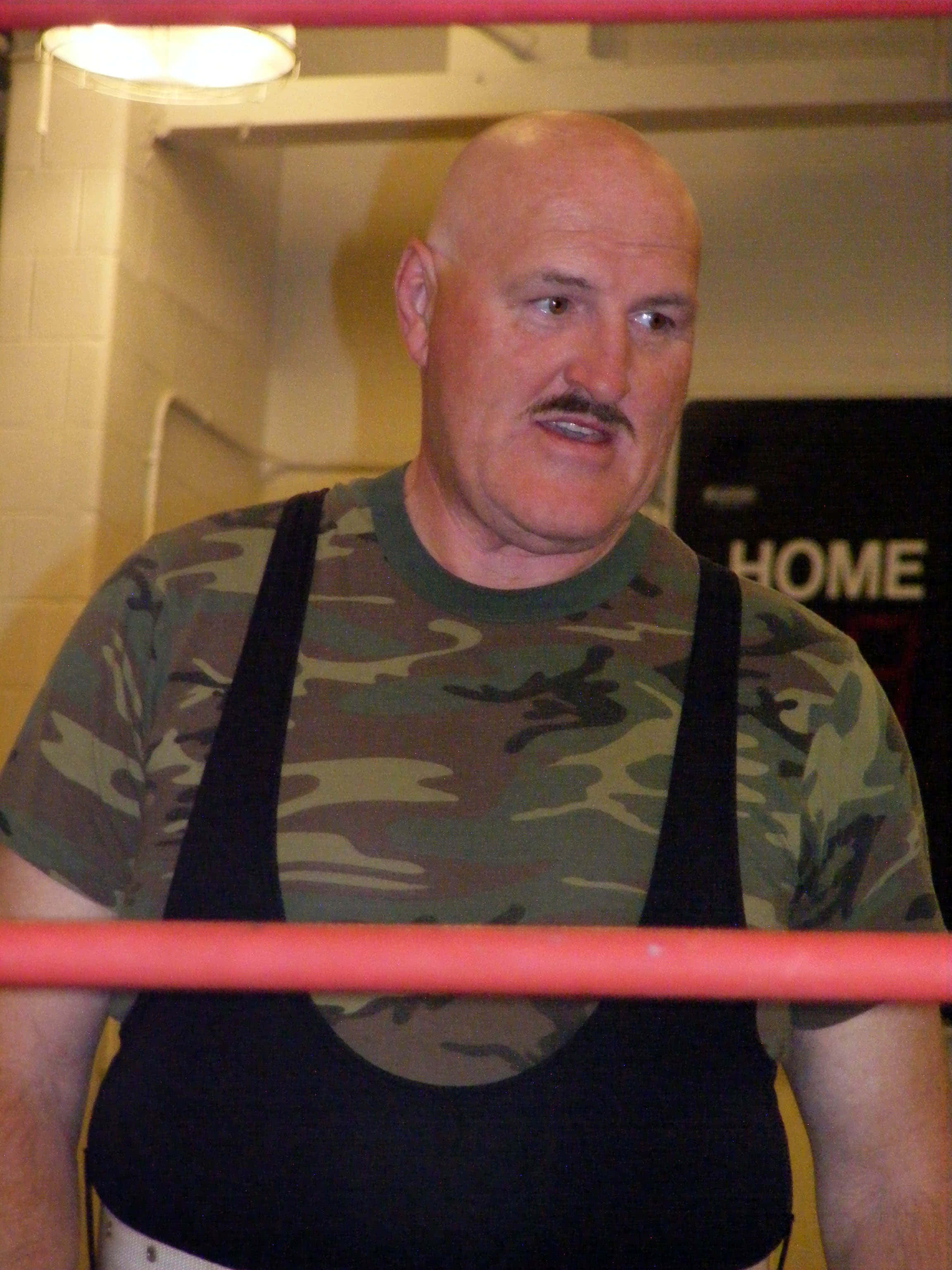 WWE 桌布 壁紙 — 一位光頭留鬍鬚的摔角手穿著迷彩襯衫，站在紅色邊框的摔角擂台內，在明亮的燈光下。