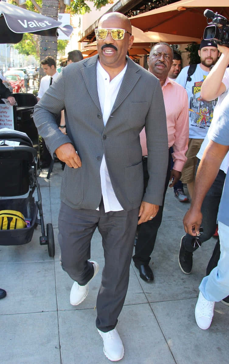 Celebrity 桌布 壁紙 — 一位光頭留鬍鬚的男人，戴著金色太陽眼鏡和灰色西裝，自信地走在陽光明媚的城市人行道上。