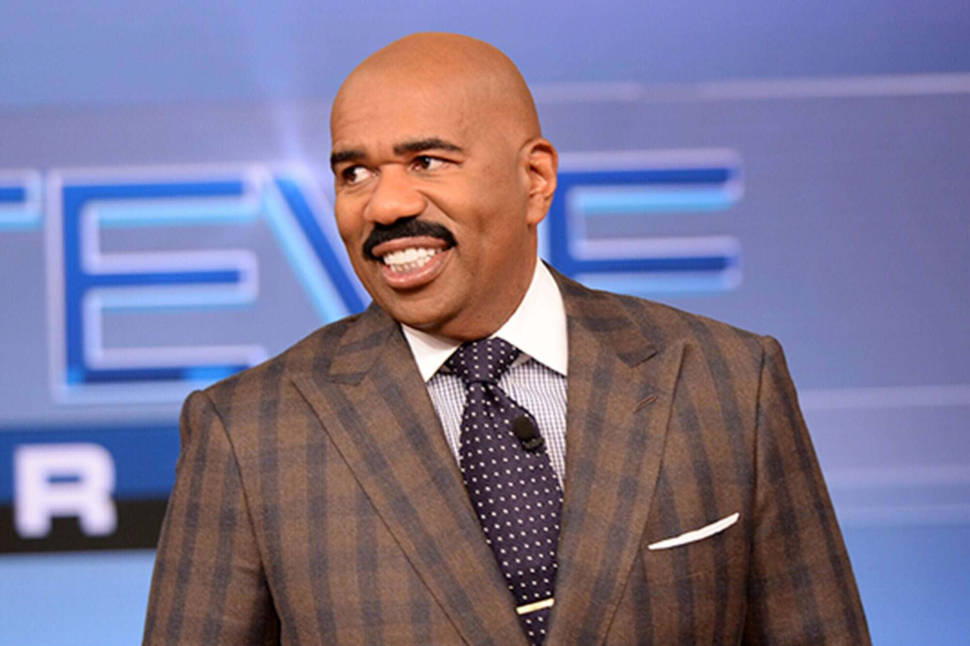 steve harvey 桌布 壁紙 — 一位光頭留鬍鬚的男人，身穿棕色格紋西裝，在藍色燈光的電視佈景中燦爛微笑。