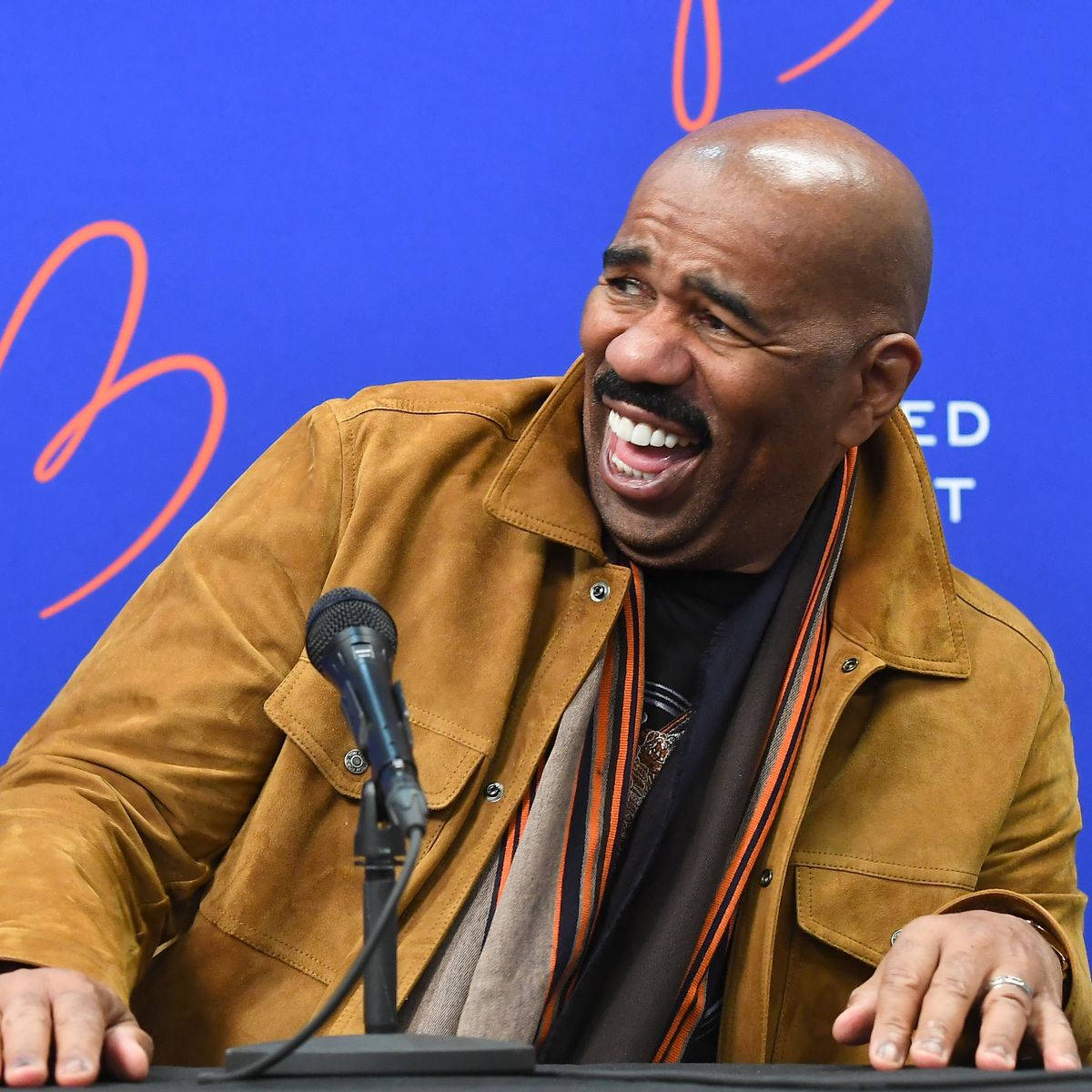 steve harvey 桌布 壁紙 — 一位光頭留鬍鬚的男人坐在講台上，對著麥克風，在藍色背景下開懷大笑。