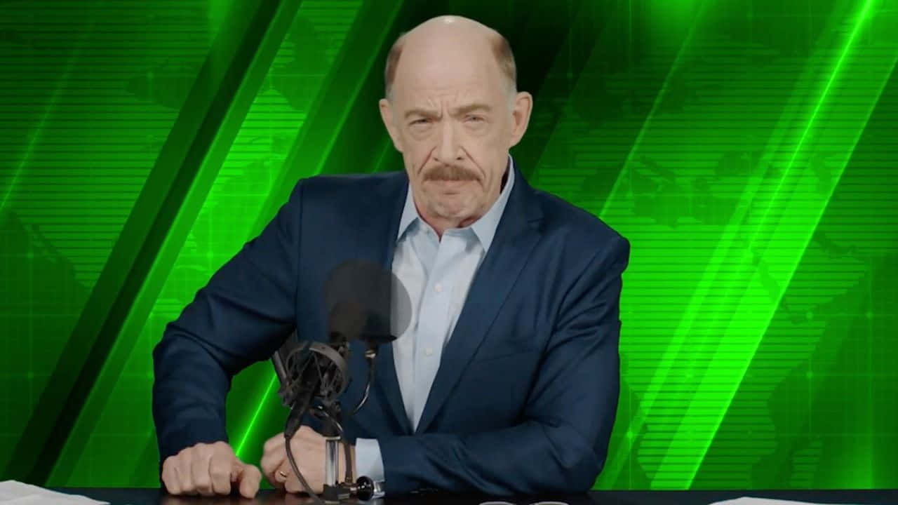 J Jonah Jameson 桌布 壁紙 — 一位光頭留鬍鬚的男人穿著藍色西裝，坐在有麥克風的桌前，背景是充滿活力的綠色數位背景。