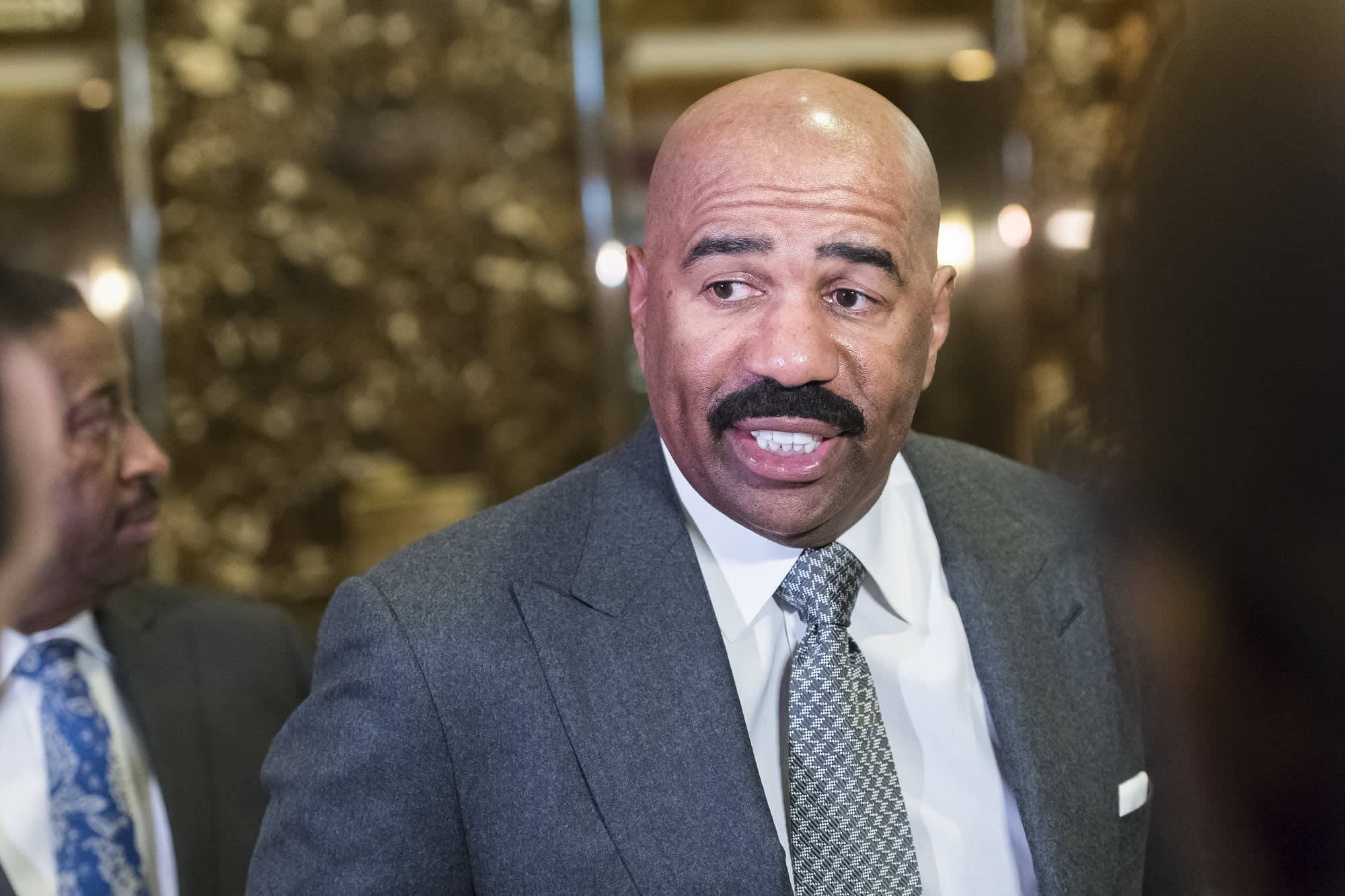 steve harvey 桌布 壁紙 — 一位光頭留鬍鬚的男士，身穿灰色西裝和花紋領帶，在室內模糊背景燈光下微笑說話。