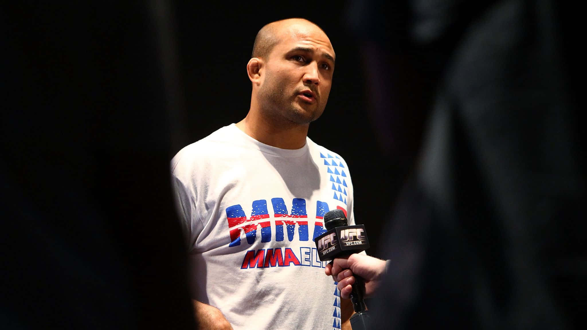 UFC 桌布 壁紙 — 一位光頭的MMA拳手，身穿帶有紅藍圖案的白襯衫，在深色背景前對著UFC麥克風說話。