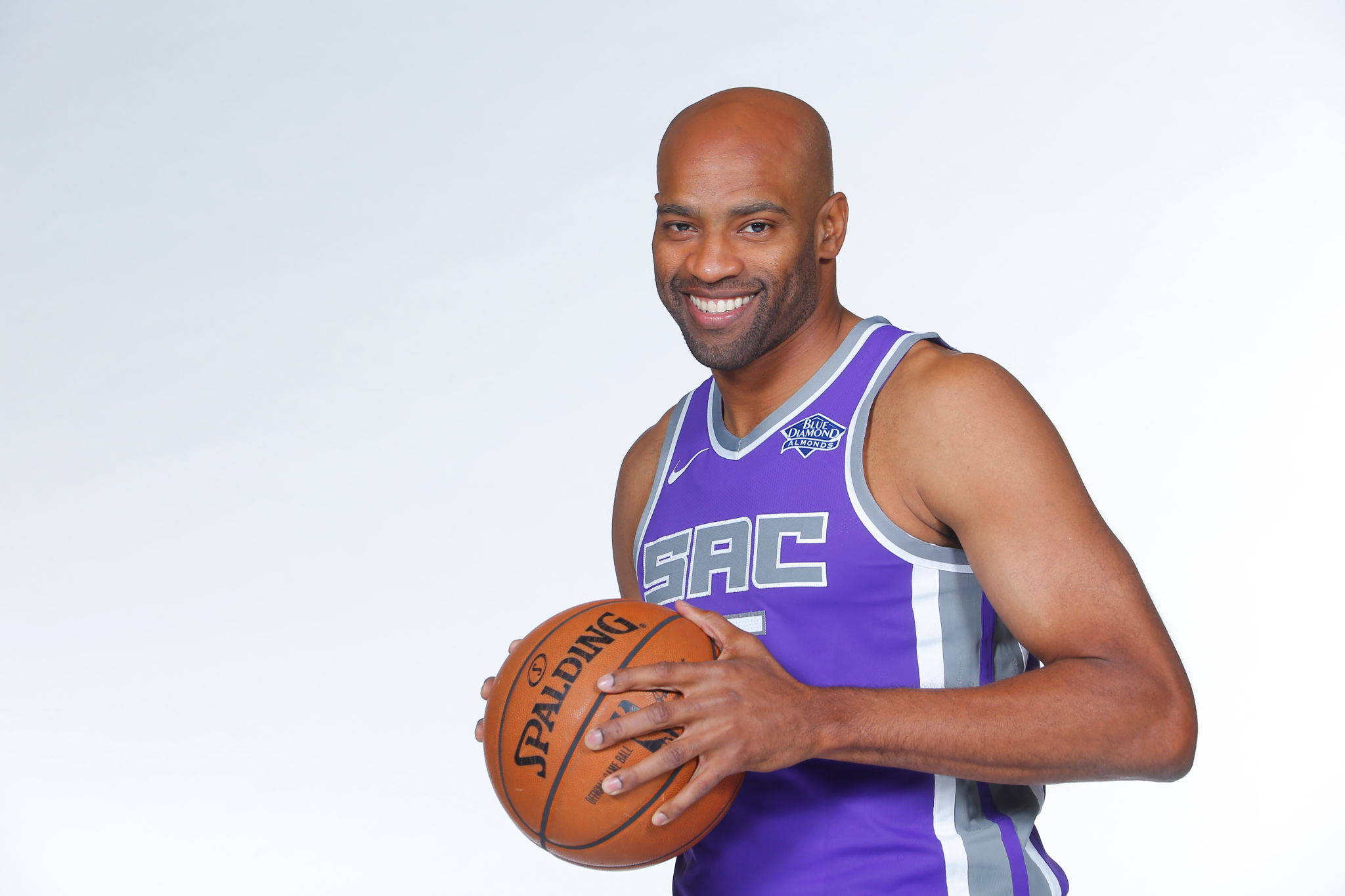 NBA 桌布 壁紙 — 一位光頭籃球員穿著紫色沙加緬度國王隊球衣，在純白背景前微笑著拿著一個Spalding籃球。
