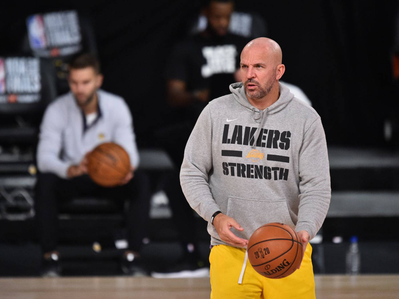 籃球 桌布 壁紙 — 一位光頭籃球教練穿著灰色 'Lakers Strength' 連帽衫和黃色短褲，在球場上拿著籃球，背景有隊友模糊的身影。