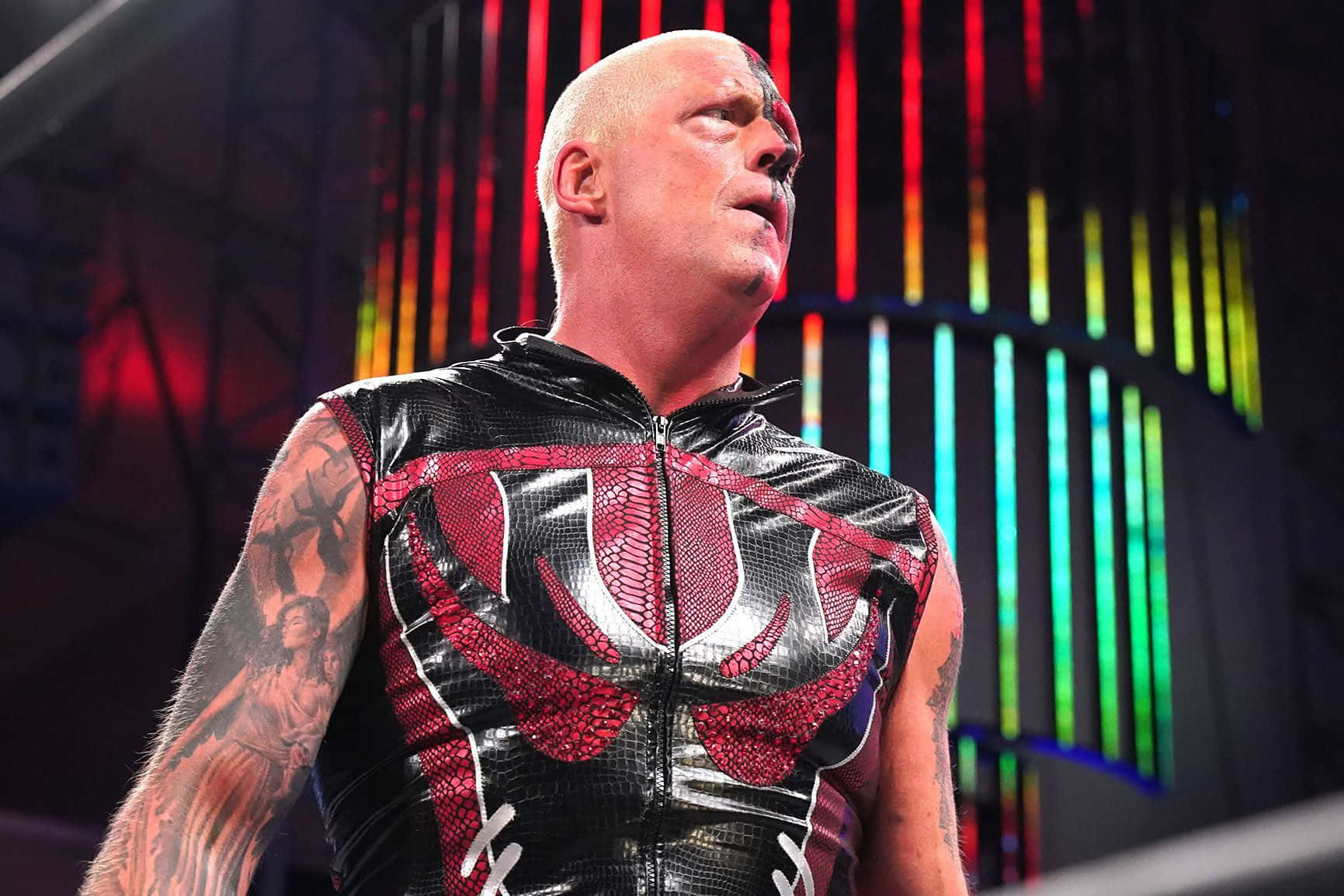 WWE 桌布 壁紙 — 一位光頭紋身摔角手穿著黑紅蛇皮背心，在彩虹燈光舞台背景下向上凝視。