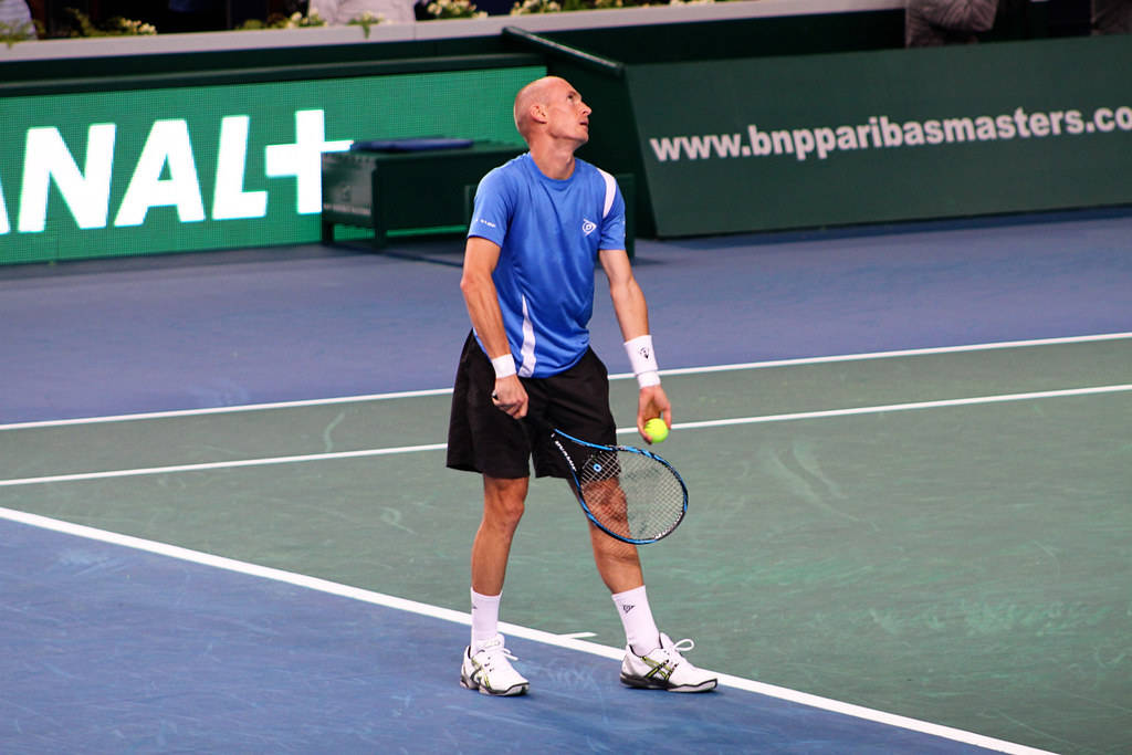 Tennis 桌布 壁紙 — 一位光頭網球運動員身穿藍色襯衫和黑色短褲，站在藍色球場上，手持球拍和球，向上看。