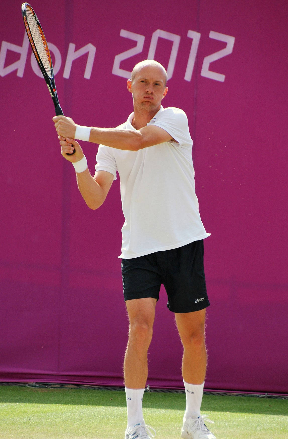 Tennis 桌布 壁紙 — 一位光頭網球選手穿著白色襯衫和黑色短褲，在草地球場上準備擊出正手拍，背景是鮮豔的紫色牆壁，上面有'London 2012'字樣。