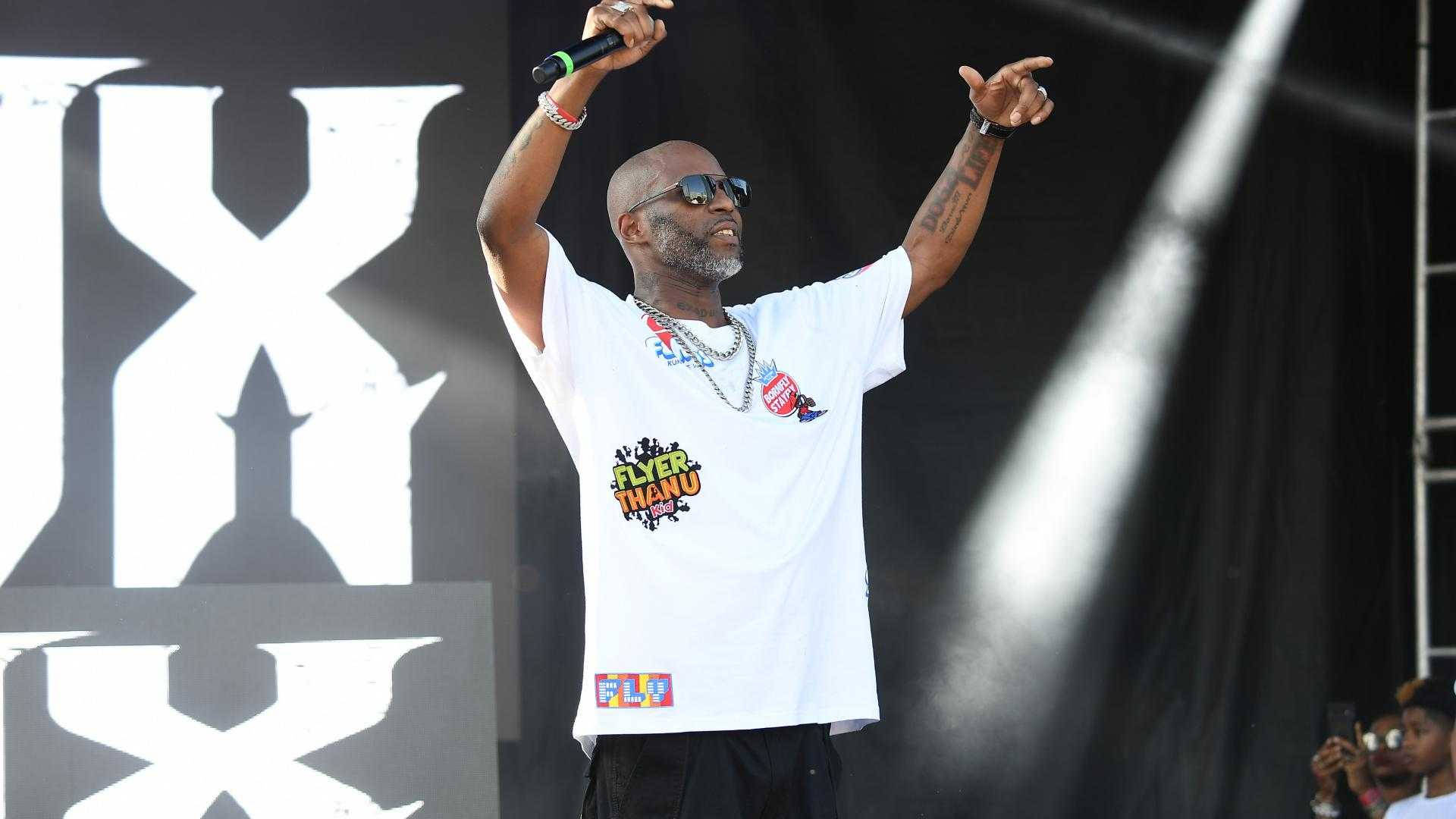 DMX 桌布 壁紙 — 一位光頭饒舌歌手戴著太陽眼鏡和白色T恤，在明亮的聚光燈下高舉雙臂，充滿活力地在舞台上表演。
