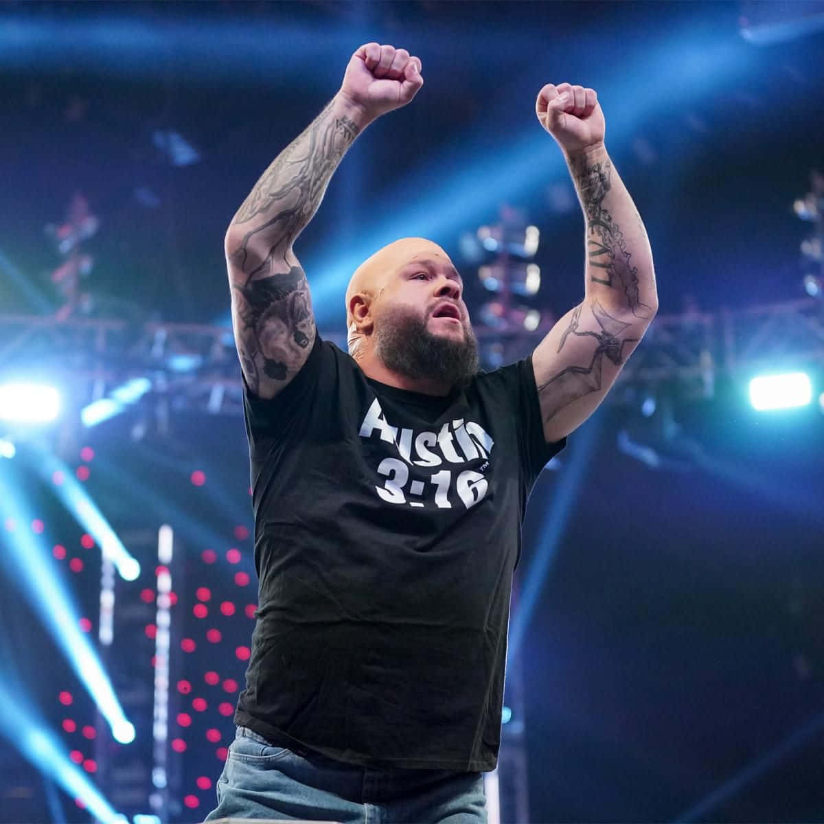 WWE 桌布 壁紙 — 一位光頭鬍鬚、手臂有刺青的摔角手在明亮的藍色舞台燈光下勝利地舉起拳頭。