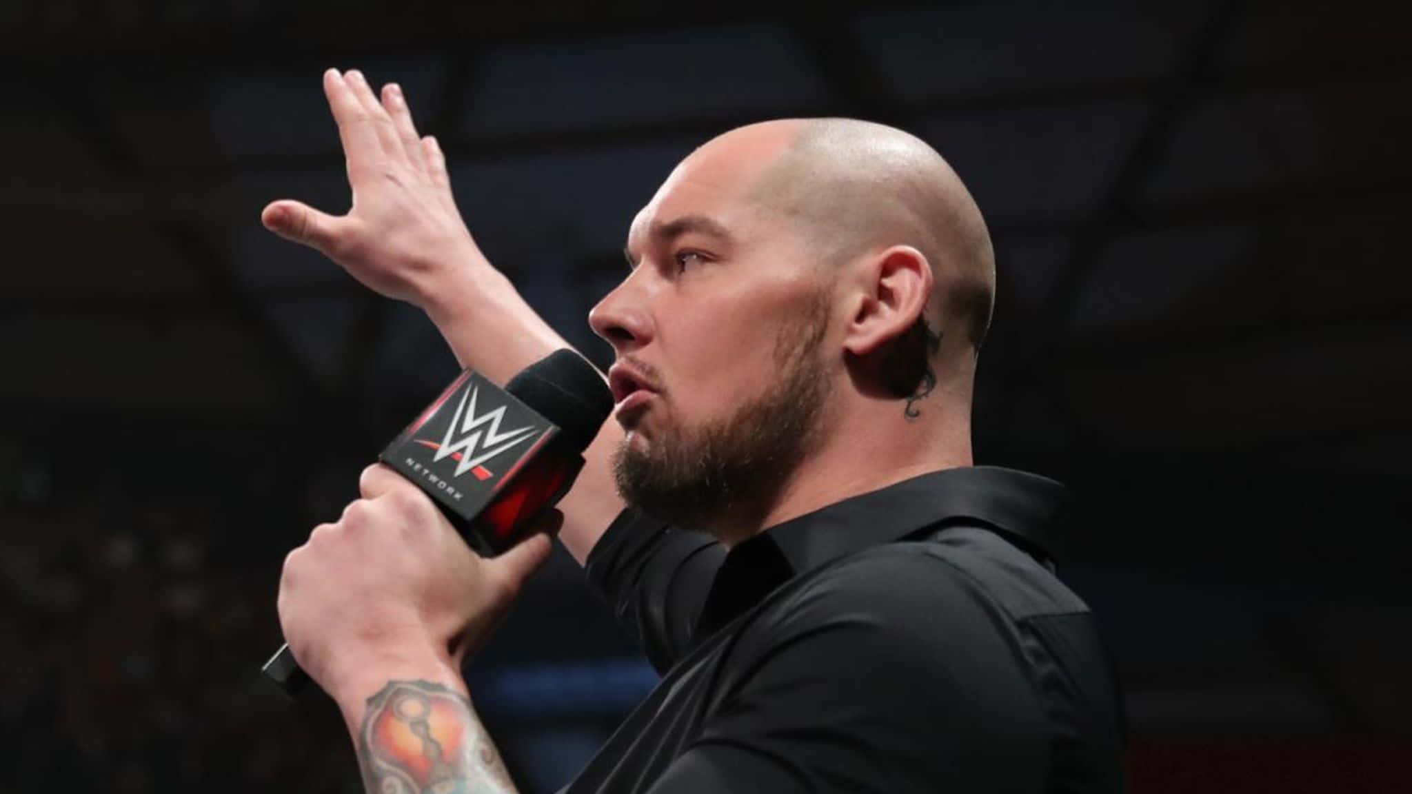 WWE 桌布 壁紙 — 一位光頭鬍鬚摔角手在明亮的舞台燈光下，於現場活動中熱情地拿著WWE麥克風並做手勢。