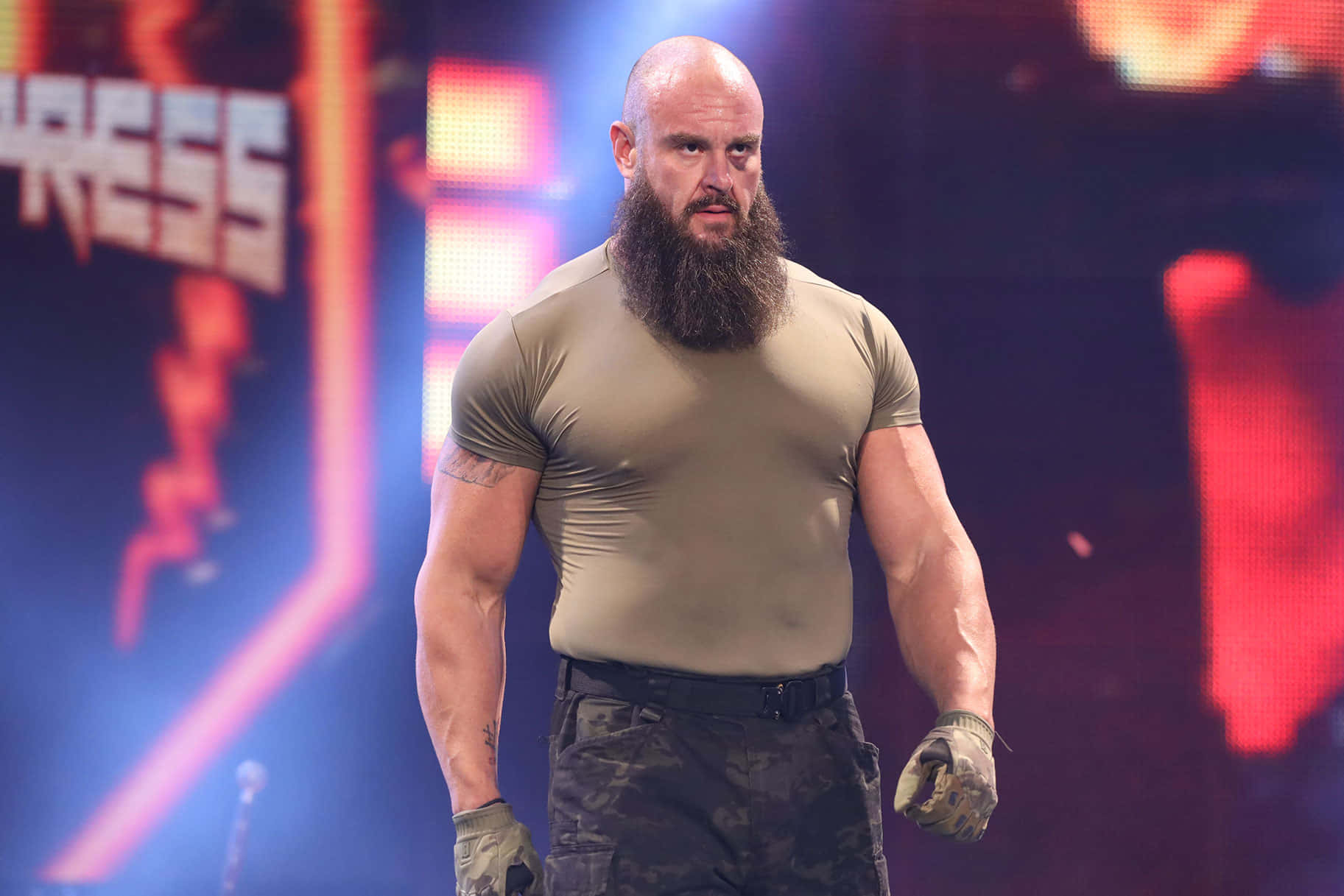 WWE 桌布 壁紙 — 一位光頭鬍鬚摔角手穿著緊身橄欖綠襯衫和迷彩褲，在充滿活力的舞台燈光下自信地大步走。