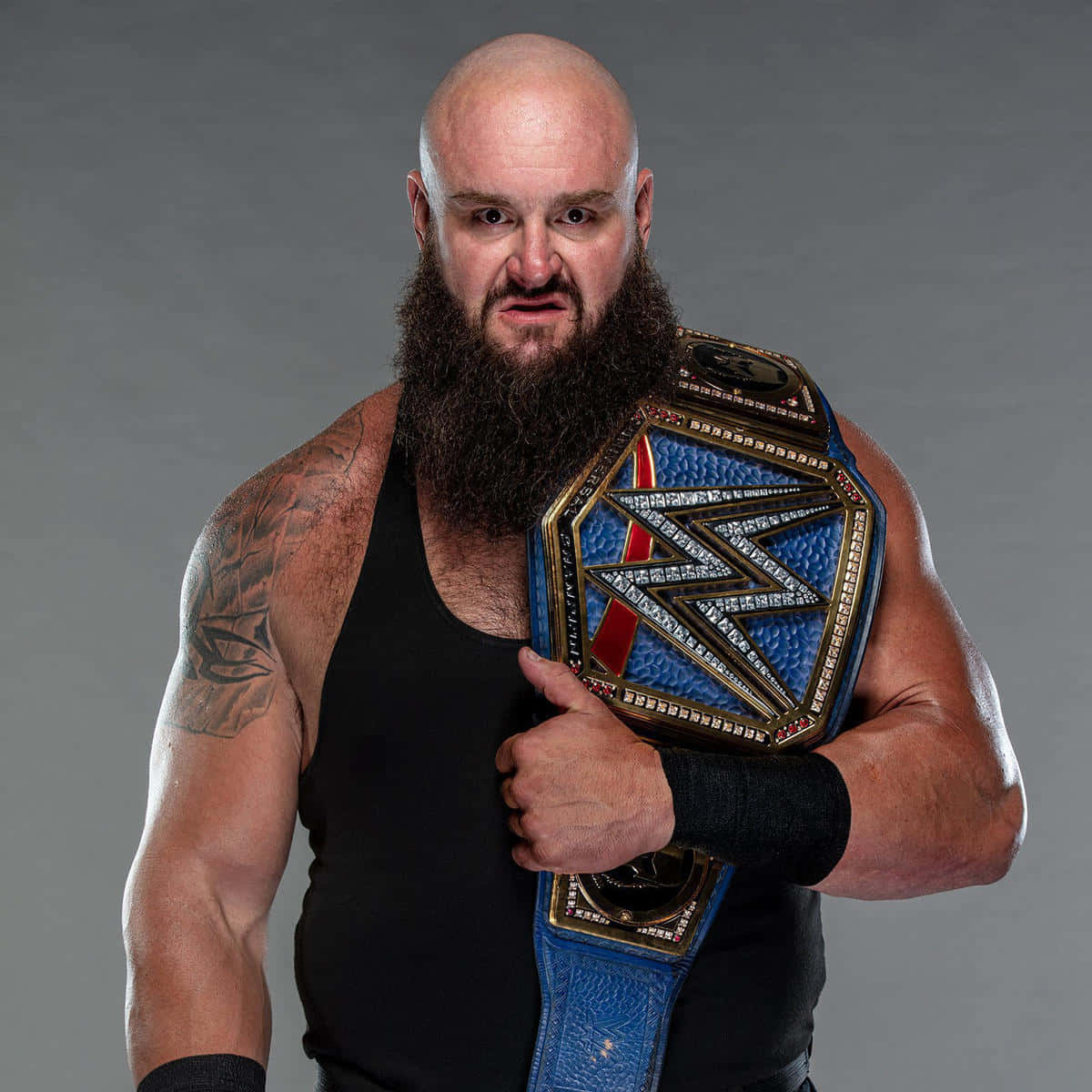 WWE 桌布 壁紙 — 一位光頭鬍鬚摔角手身穿黑色背心，在灰色攝影棚背景下緊握一條藍金色冠軍腰帶。