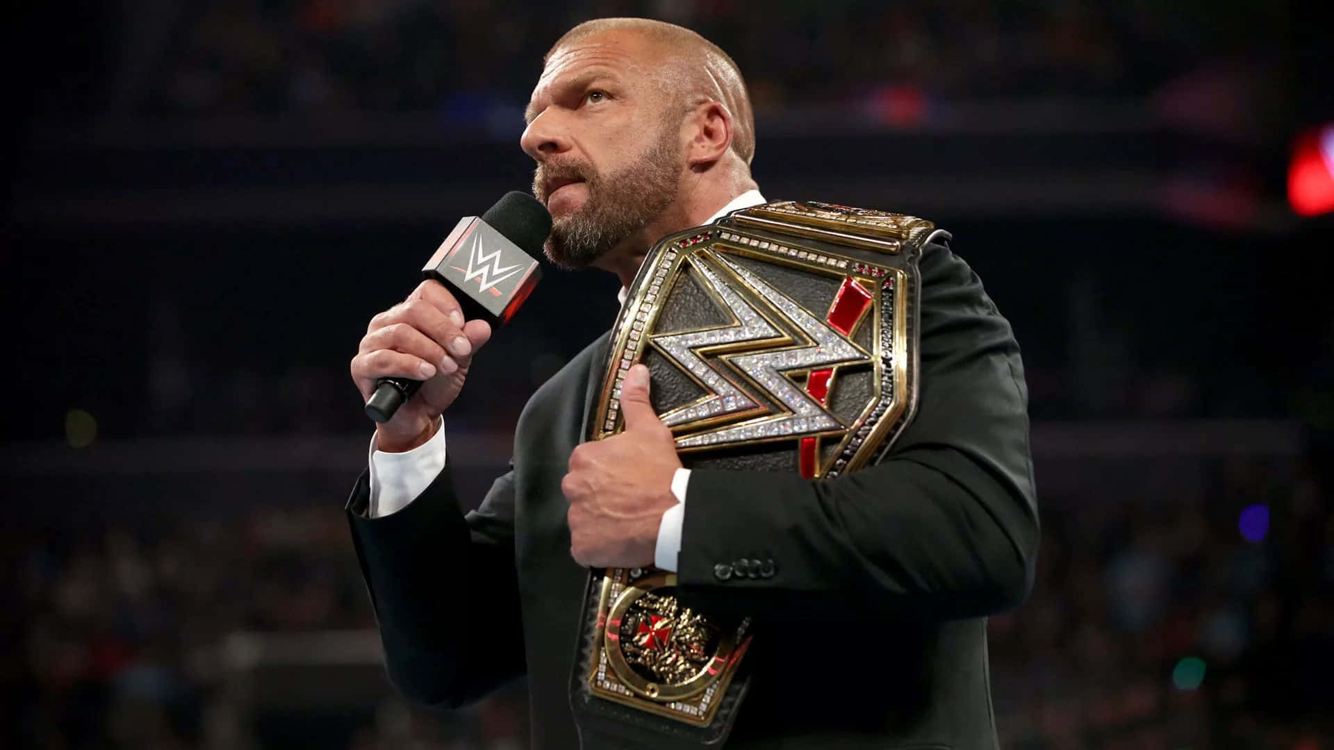WWE 桌布 壁紙 — 一位光頭鬍鬚摔角手身穿黑色西裝，在昏暗的場館中手持WWE冠軍腰帶和麥克風向人群講話。