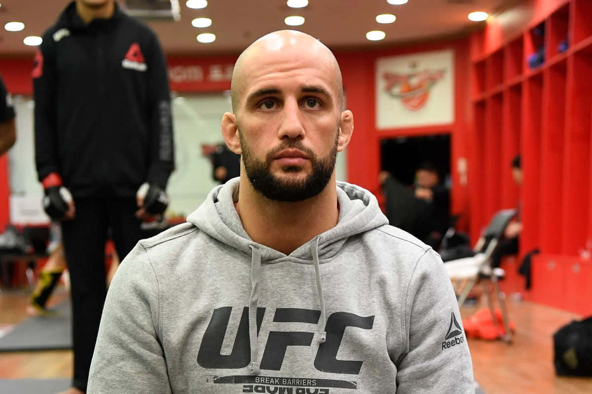 MMA 桌布 壁紙 — 一位光頭鬍鬚的戰士穿著灰色UFC連帽衫，在紅色燈光的更衣室中凝視著。