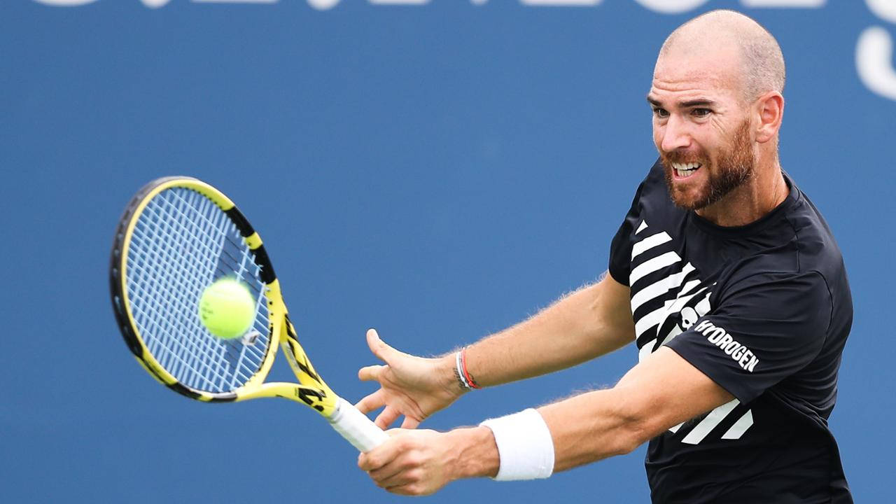US Open 桌布 壁紙 — 一位光頭鬍鬚的網球選手身穿黑白條紋，揮動黃色球拍擊打亮綠色球，背景為純藍色。