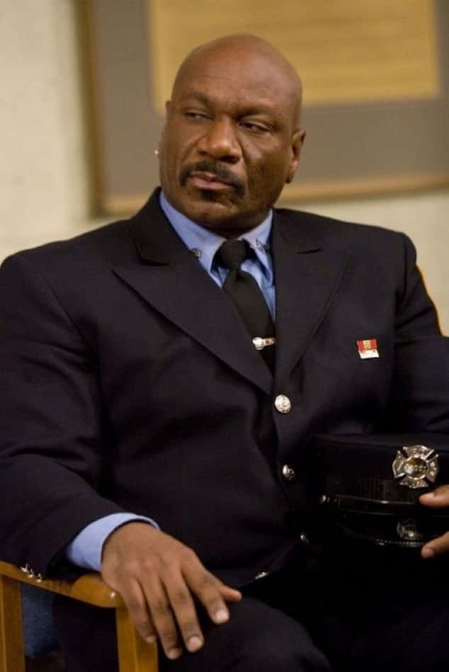 Ving Rhames 桌布 壁紙 — 一位光頭黑人男子身穿深色消防員制服，冷靜地坐著，手持帽子，表情嚴肅。