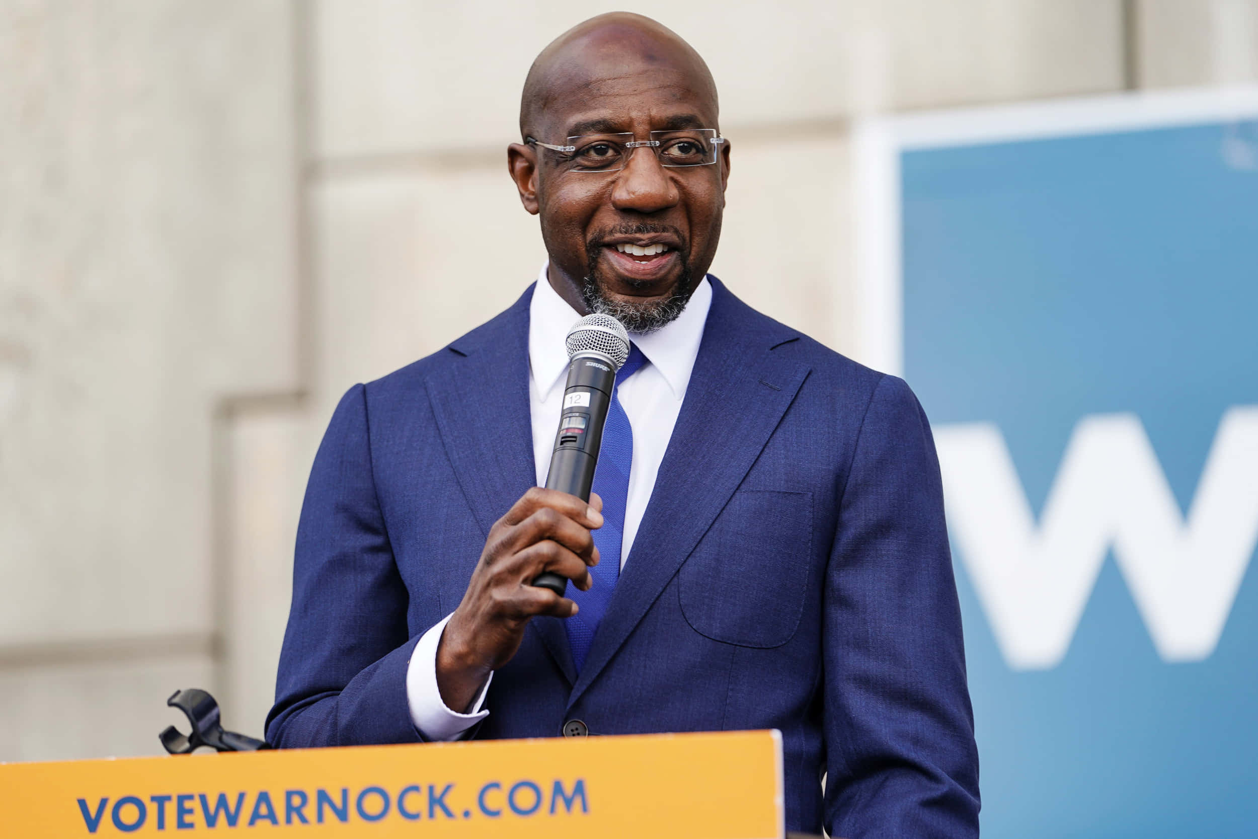 桌布 壁紙 — 一位光頭黑人男子身穿藍色西裝戴眼鏡，在設有「VOTEWARNOCK.COM」講台的競選活動中對著麥克風發言。