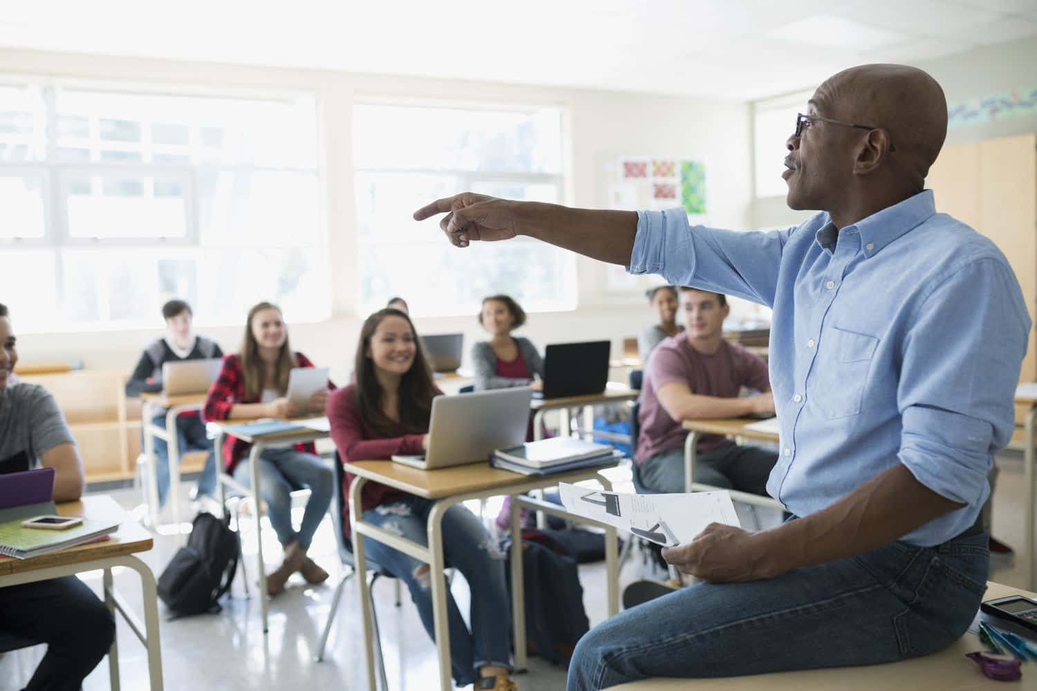 High School 桌布 壁紙 — 一位光頭黑人男教師穿著藍色襯衫，權威地指向坐在明亮教室書桌前的學生。