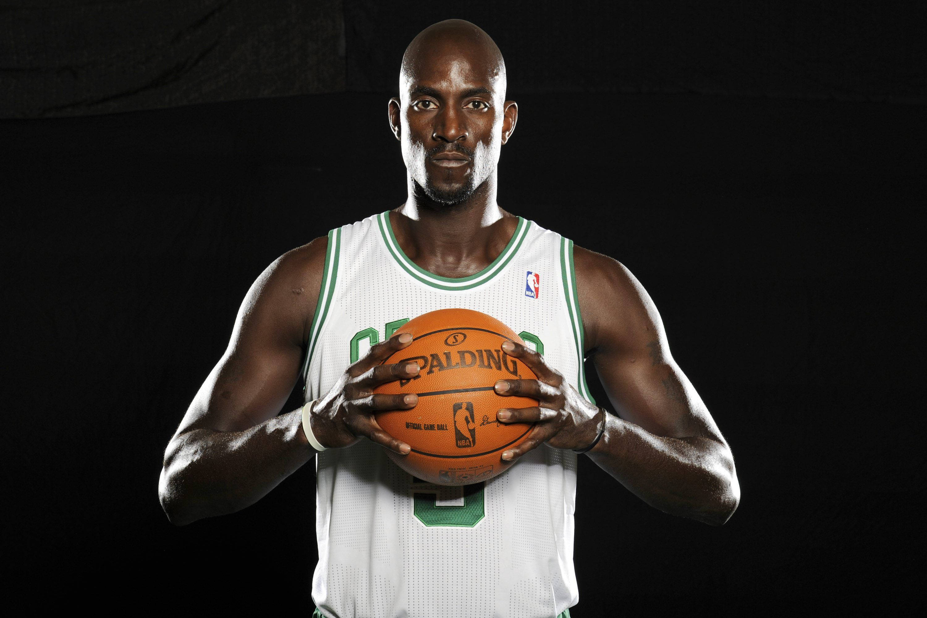 Kevin Garnett 桌布 壁紙 — 一位光頭黑人籃球運動員身穿白色和綠色球衣，在黑色背景前拿著一個橙色 Spalding 籃球。