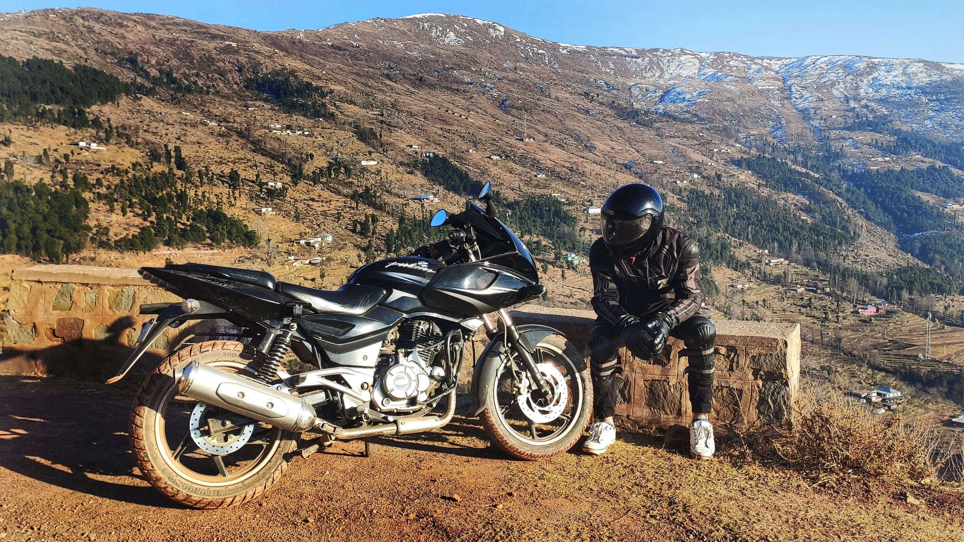 Pulsar 220 HD 桌布 壁紙 — 一位全副裝備的摩托車手坐在他們的黑色摩托車旁邊，在一個石頭邊緣上，俯瞰著晴朗藍天下積雪的山脈。