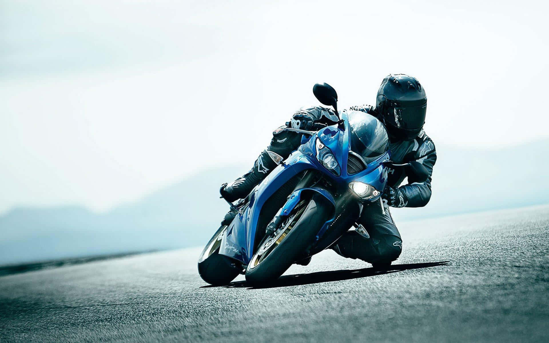 機車 桌布 壁紙 — 一位全副裝備的騎士在陽光明媚的開闊道路上，騎著流線型藍色運動機車傾斜入彎。