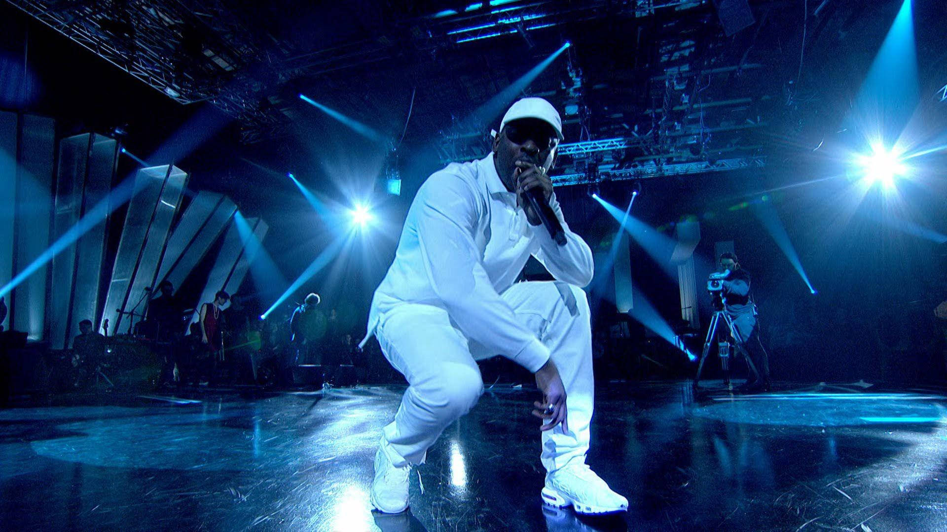 Skepta 桌布 壁紙 — 一位全白裝扮的饒舌歌手在明亮的藍色聚光燈下蹲在舞台中央，身後有樂隊。