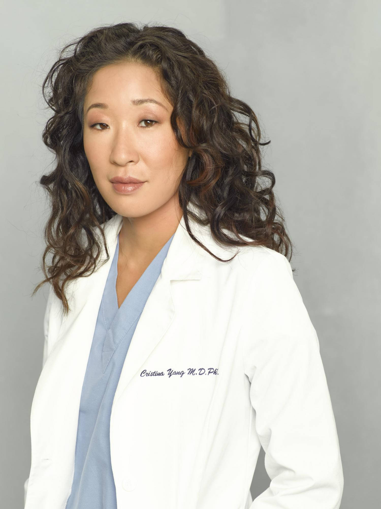 greys anatomy 桌布 壁紙 — 一位冷靜的女性醫生，留著捲曲的深色頭髮，身穿藍色刷手服外罩白色實驗室外套，背景為中性灰色。