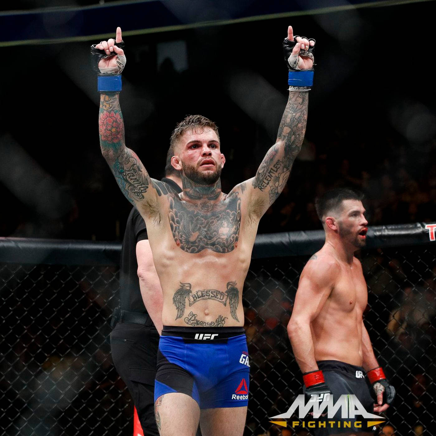 桌布 壁紙 — 一位刺青MMA拳手在昏暗的八角籠內，高舉雙臂慶祝勝利，旁邊是被擊敗的對手。