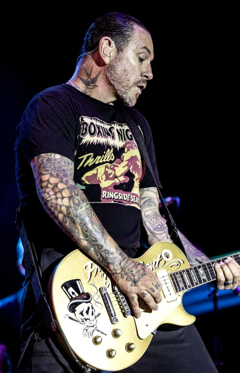 guitar player 桌布 壁紙 — 一位刺青吉他手在現場音樂會表演期間，在舞台燈光下彈奏金色電吉他。
