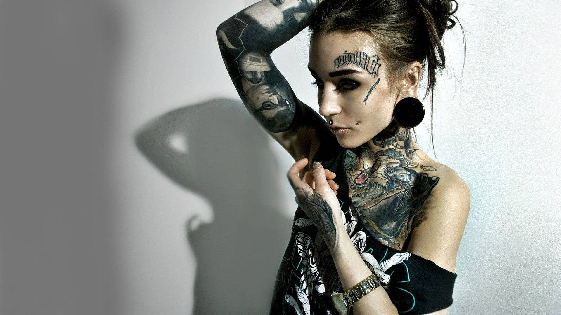 Tattoo 桌布 壁紙 — 一位刺青女子，化著深色妝容，戴著大圈耳環，靠著素色牆壁擺姿勢，投射出戲劇性的陰影。