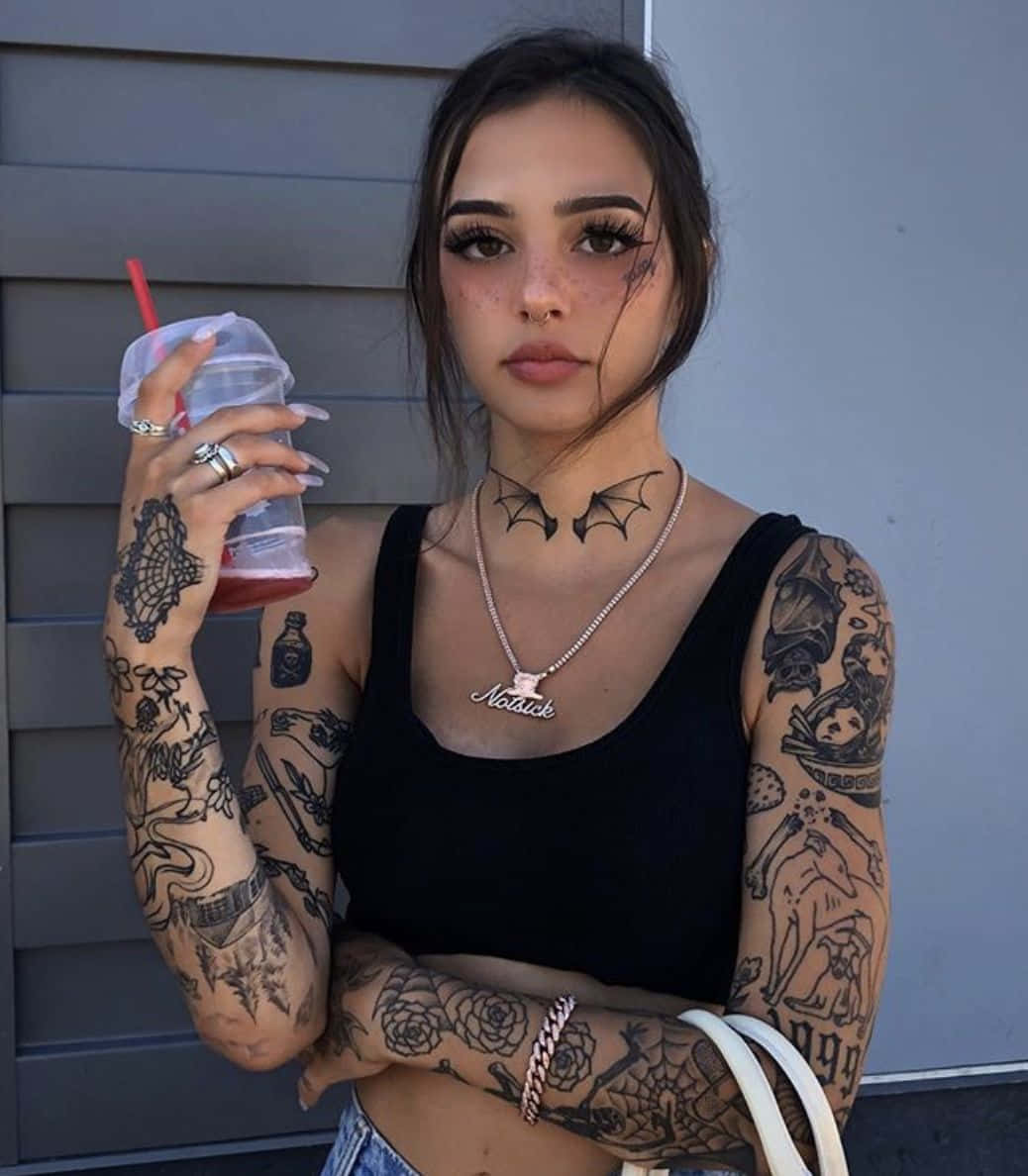 Tattoo Girl 桌布 壁紙 — 一位刺青女子戴著蝙蝠項鍊，手持飲料，靠著灰色牆壁，眼神自信。