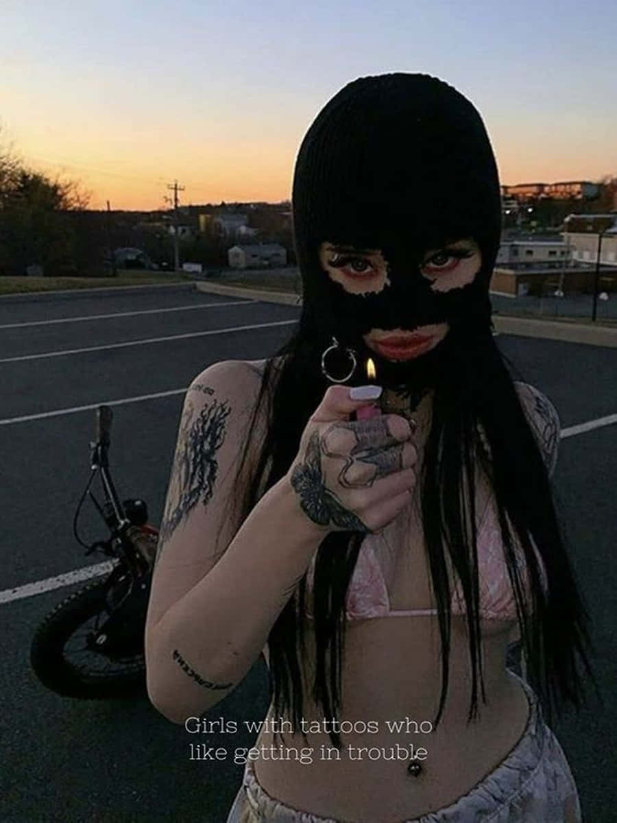 Ski Mask Girl 桌布 壁紙 — 一位刺青女子戴著黑色巴拉克拉法帽，在日落時分的停車場拿著點燃的打火機。