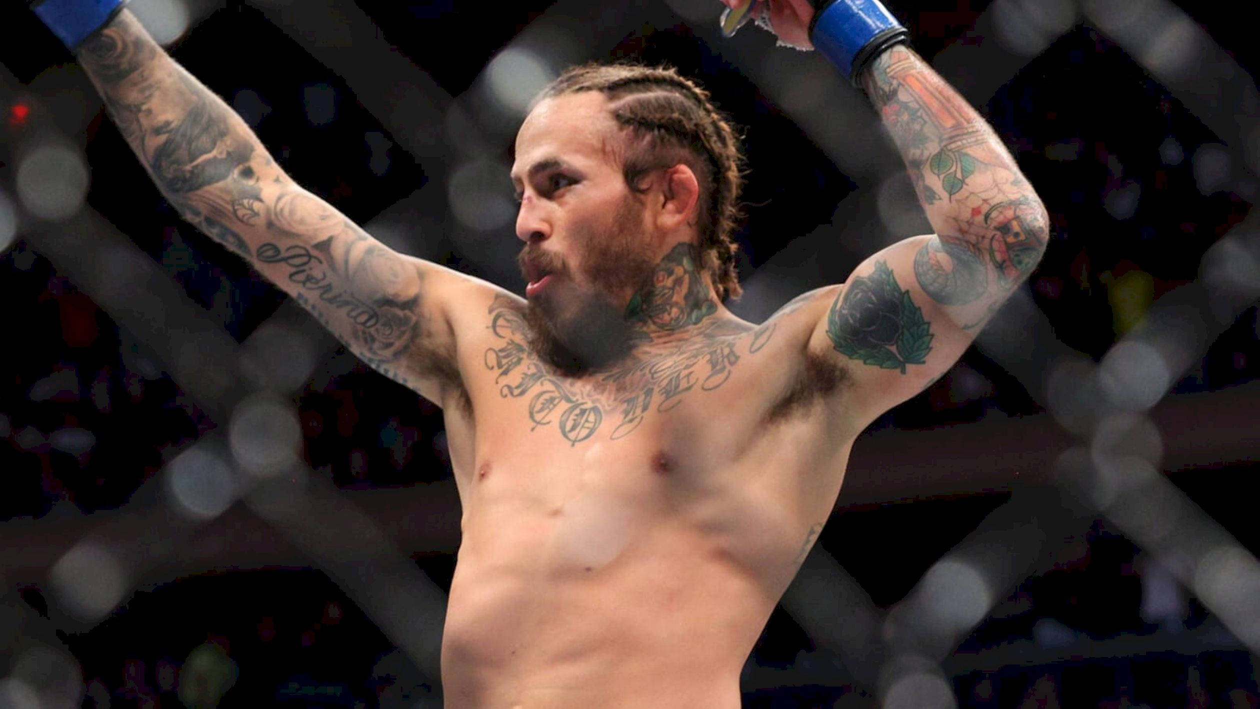 UFC 桌布 壁紙 — 一位刺青混合武術家在明亮的場館燈光下於八角籠中高舉雙臂慶祝勝利。
