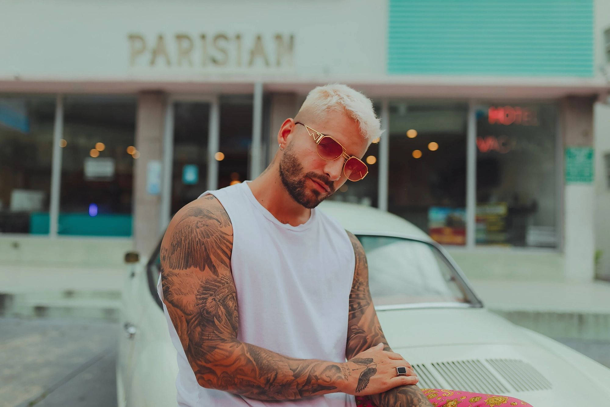 Maluma 桌布 壁紙 — 一位刺青男子，漂染金髮，戴著金框太陽眼鏡，在巴黎一間飯店前靠著一輛白色經典汽車。