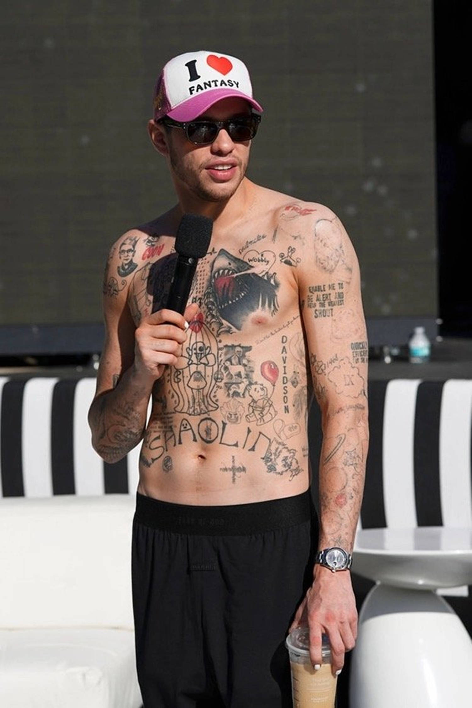 Pete Davidson 桌布 壁紙 — 一位刺青男子戴著粉白色帽子和太陽眼鏡，站在黑白條紋家具前，手持麥克風和飲料。