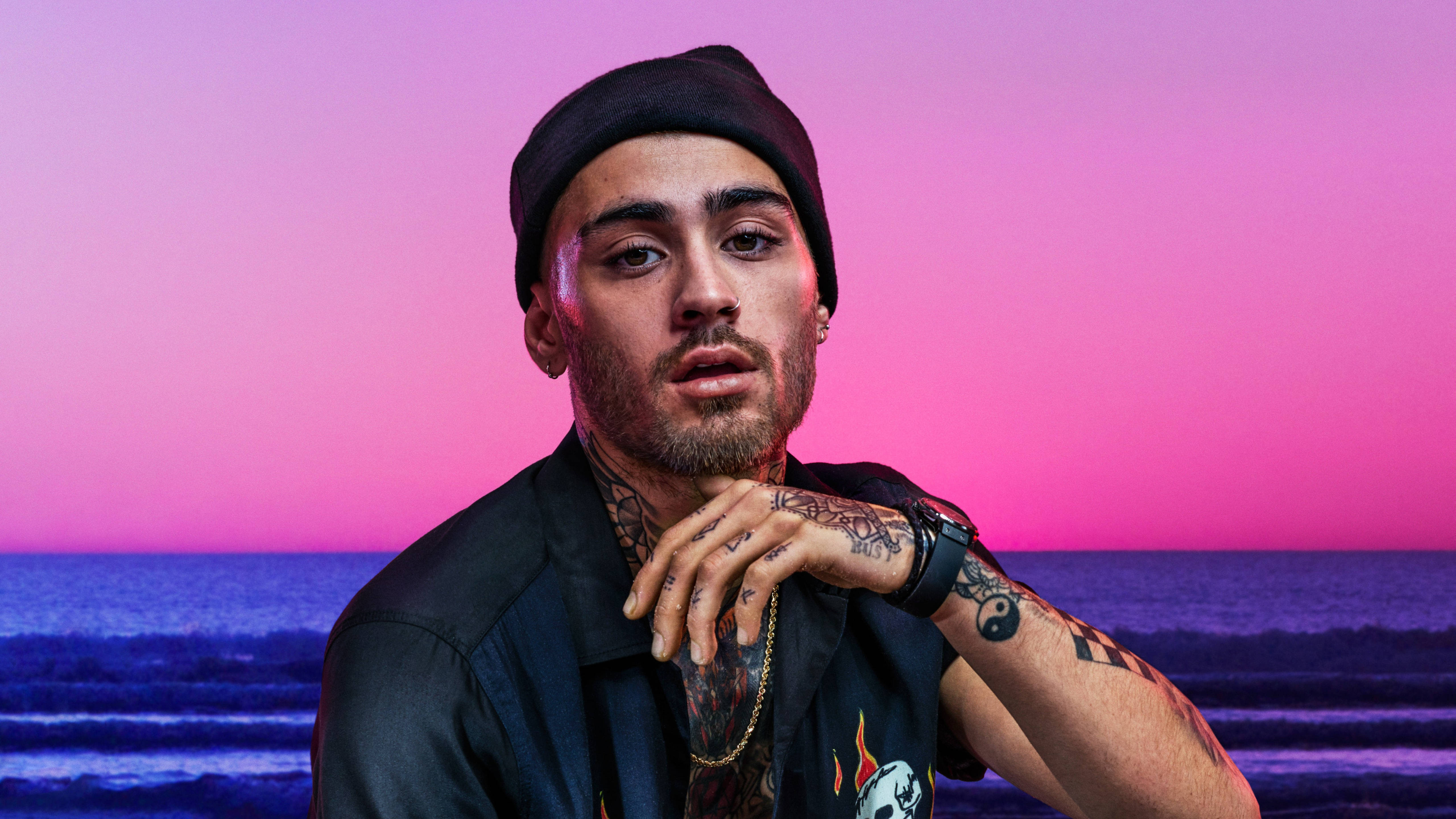 zayn 桌布 壁紙 — 一位刺青男子戴著黑色毛帽，在海洋上充滿活力的粉紅色和紫色日落前擺姿勢。