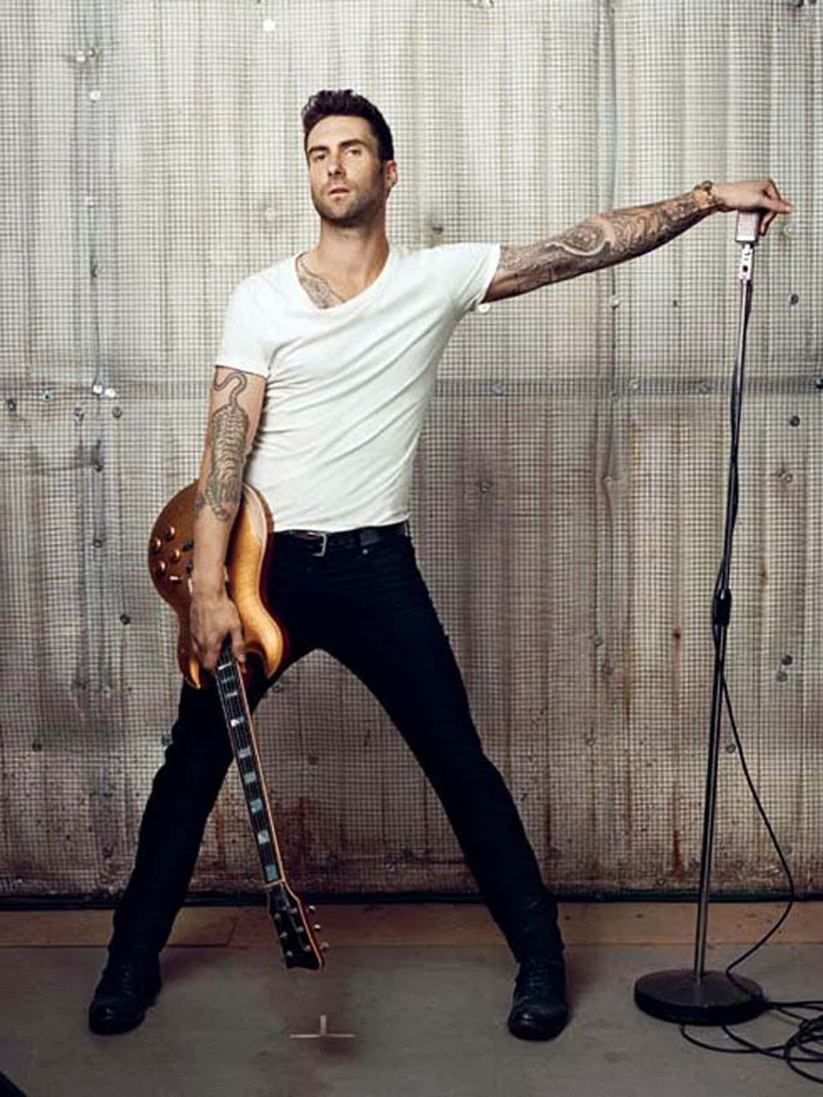 Adam Levine 桌布 壁紙 — 一位刺青男子身穿白色T恤和黑色長褲，在生鏽的金屬牆前自信地擺姿勢，手持吉他和麥克風。