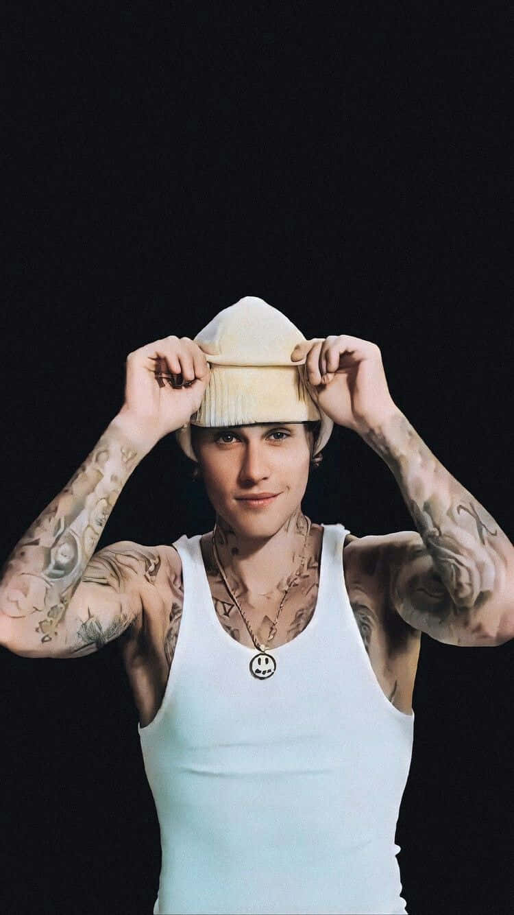Justin Bieber 2021 桌布 壁紙 — 一位刺青男子身穿白色背心和米色無邊帽，在純黑背景下調整帽子。