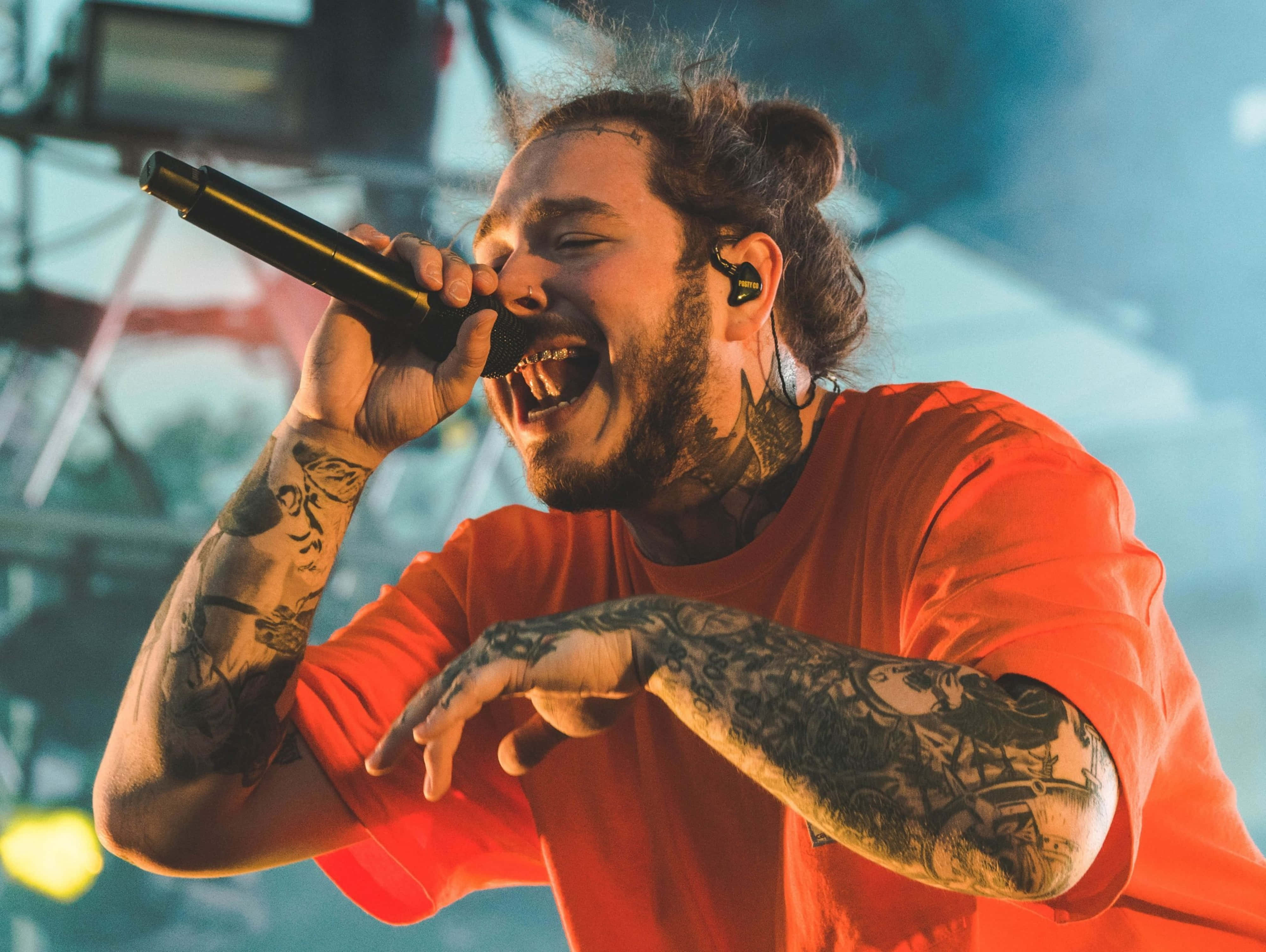 4K Rapper 桌布 壁紙 — 一位刺青男歌手穿著紅色襯衫，在明亮的燈光下於音樂會舞台上熱情地對著麥克風演唱。