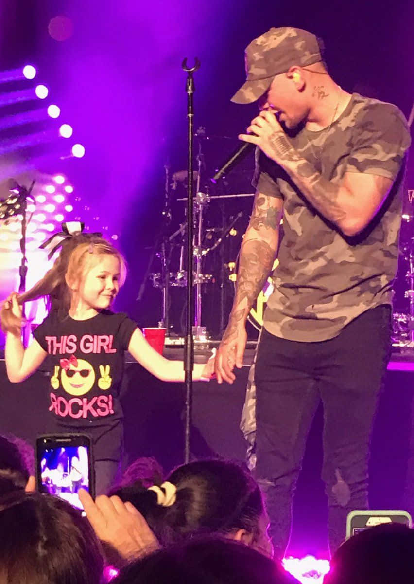 Kane Brown 桌布 壁紙 — 一位刺青男歌手身穿迷彩服，在紫色舞台燈光下的演唱會中與微笑的年輕女孩牽手。