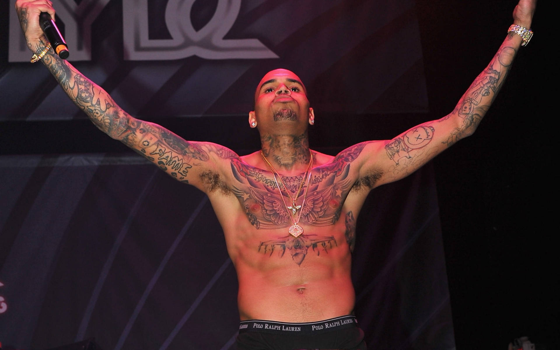 Chris Brown 桌布 壁紙 — 一位刺青男表演者在紅燈下舞台上高舉雙臂，手持麥克風，展現勝利姿態。