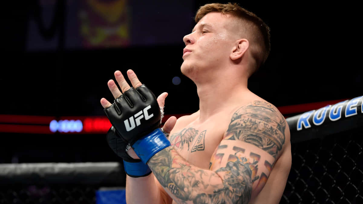UFC 桌布 壁紙 — 一位刺青的UFC拳手戴著黑色手套，在明亮的場館燈光下於八角籠內高舉雙手慶祝勝利。