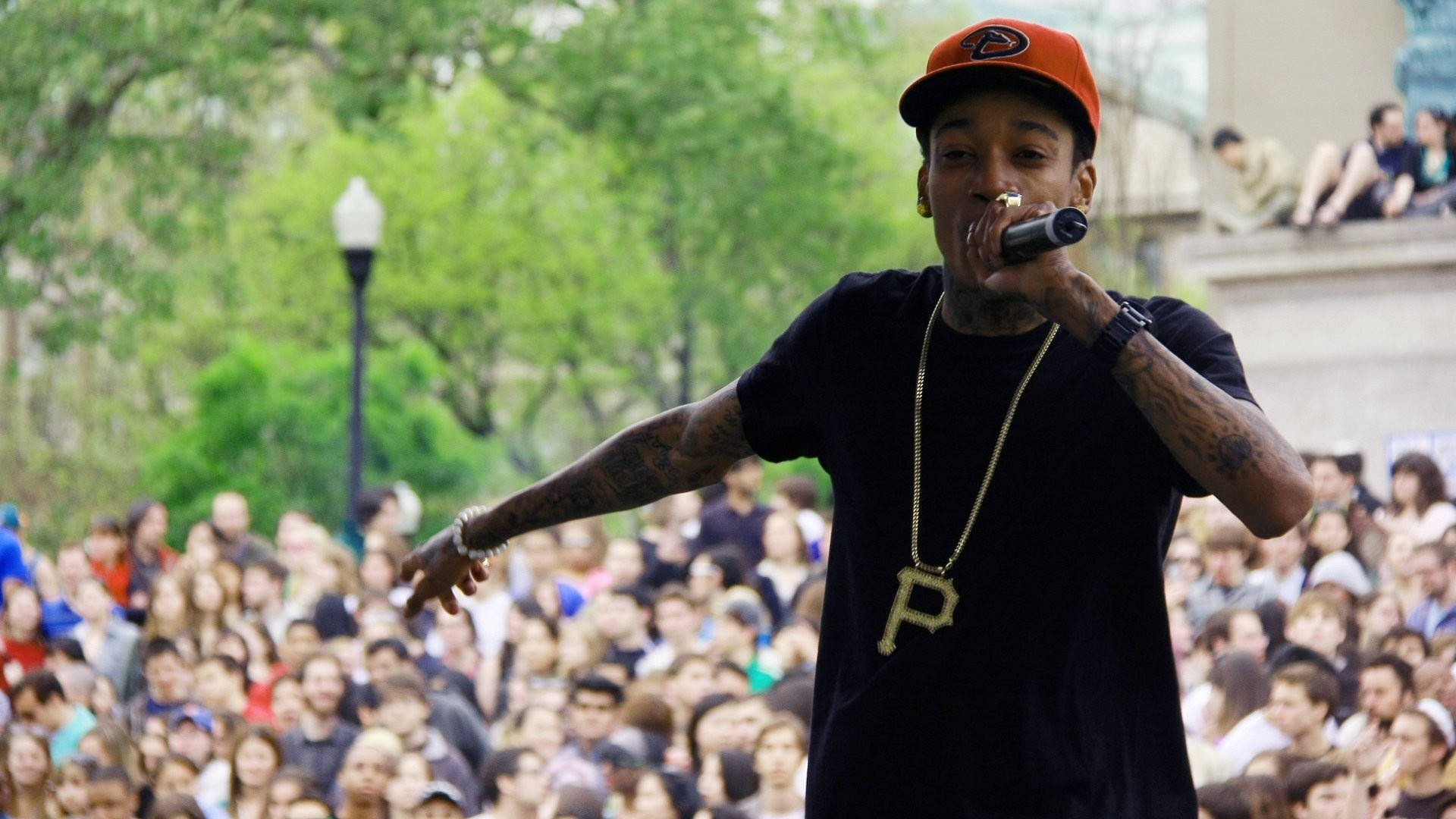 wiz khalifa 桌布 壁紙 — 一位刺青說唱歌手戴著橘色帽子和黑色襯衫，在綠色樹木下的大型戶外人群前用麥克風表演。
