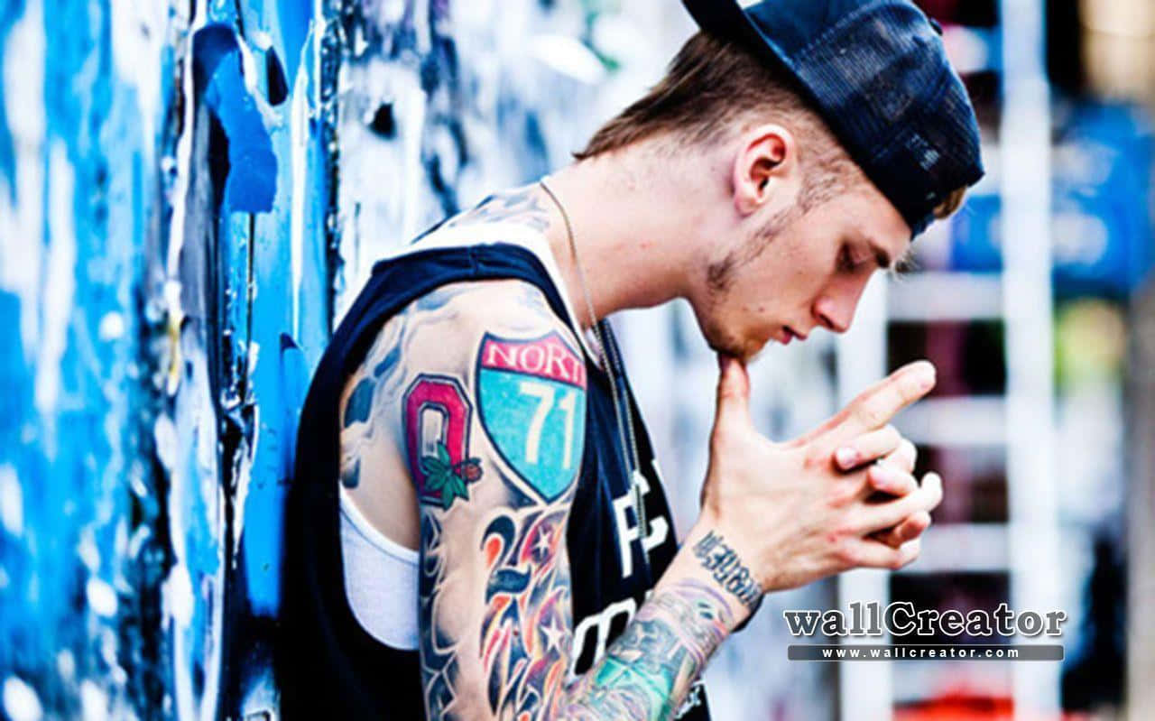mgk 桌布 壁紙 — 一位刺青青年戴著帽子，靠在一面佈滿塗鴉的藍色牆上，手指輕觸嘴唇，擺出沉思的姿勢。