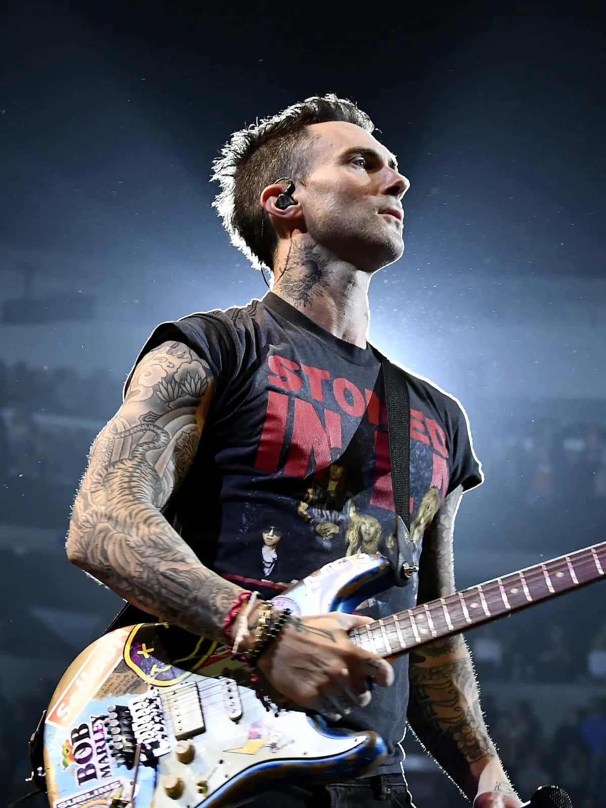 Adam Levine 桌布 壁紙 — 一位刺青音樂家在明亮的舞台燈光下於現場音樂會表演中彈奏電吉他。