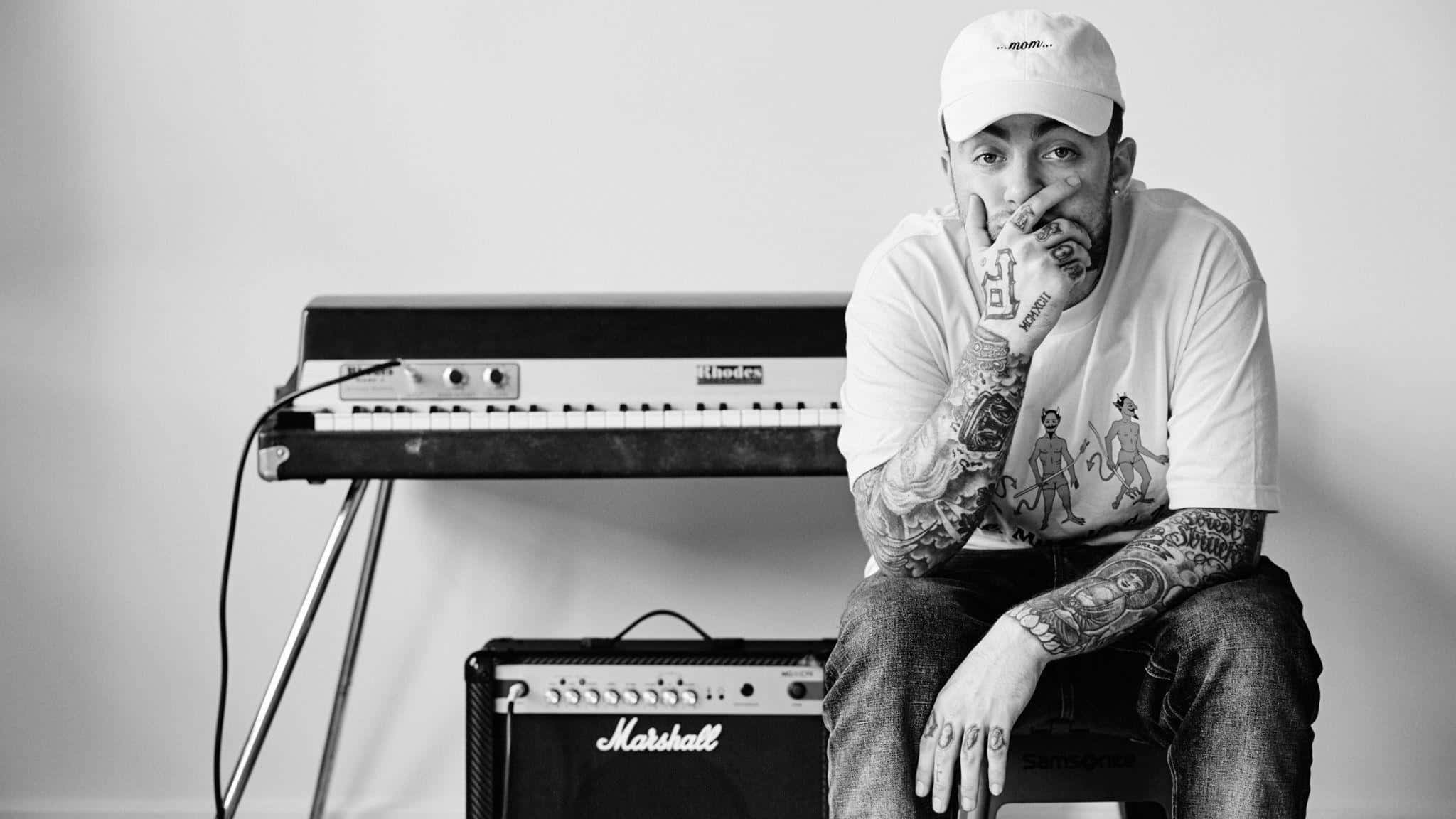 Mac Miller 桌布 壁紙 — 一位刺青音樂家戴著白色帽子，在極簡黑白工作室中若有所思地坐在鍵盤和 Marshall 擴音器旁邊。